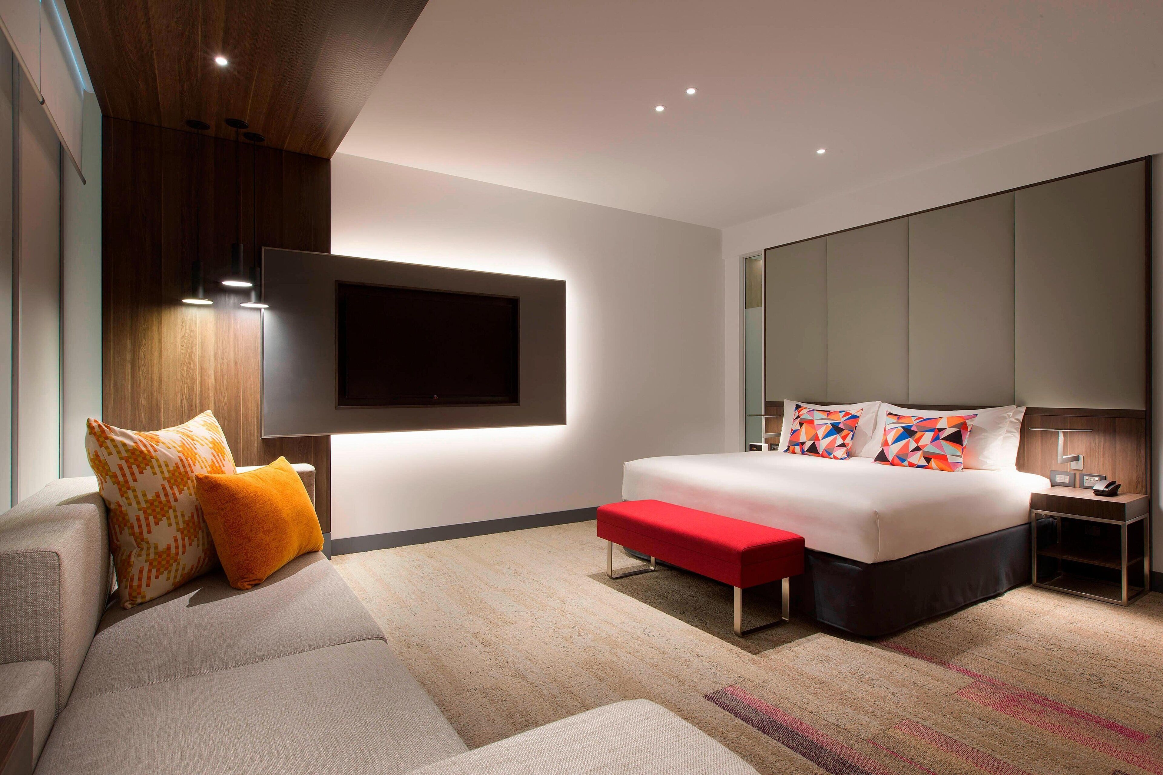 Un dormitorio moderno con una cama grande, un televisor montado en la pared, un banco rojo y una mesita de noche.