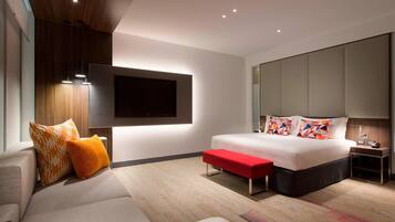 Un dormitorio moderno con una cama grande, un televisor montado en la pared, un banco rojo y una mesita de noche.