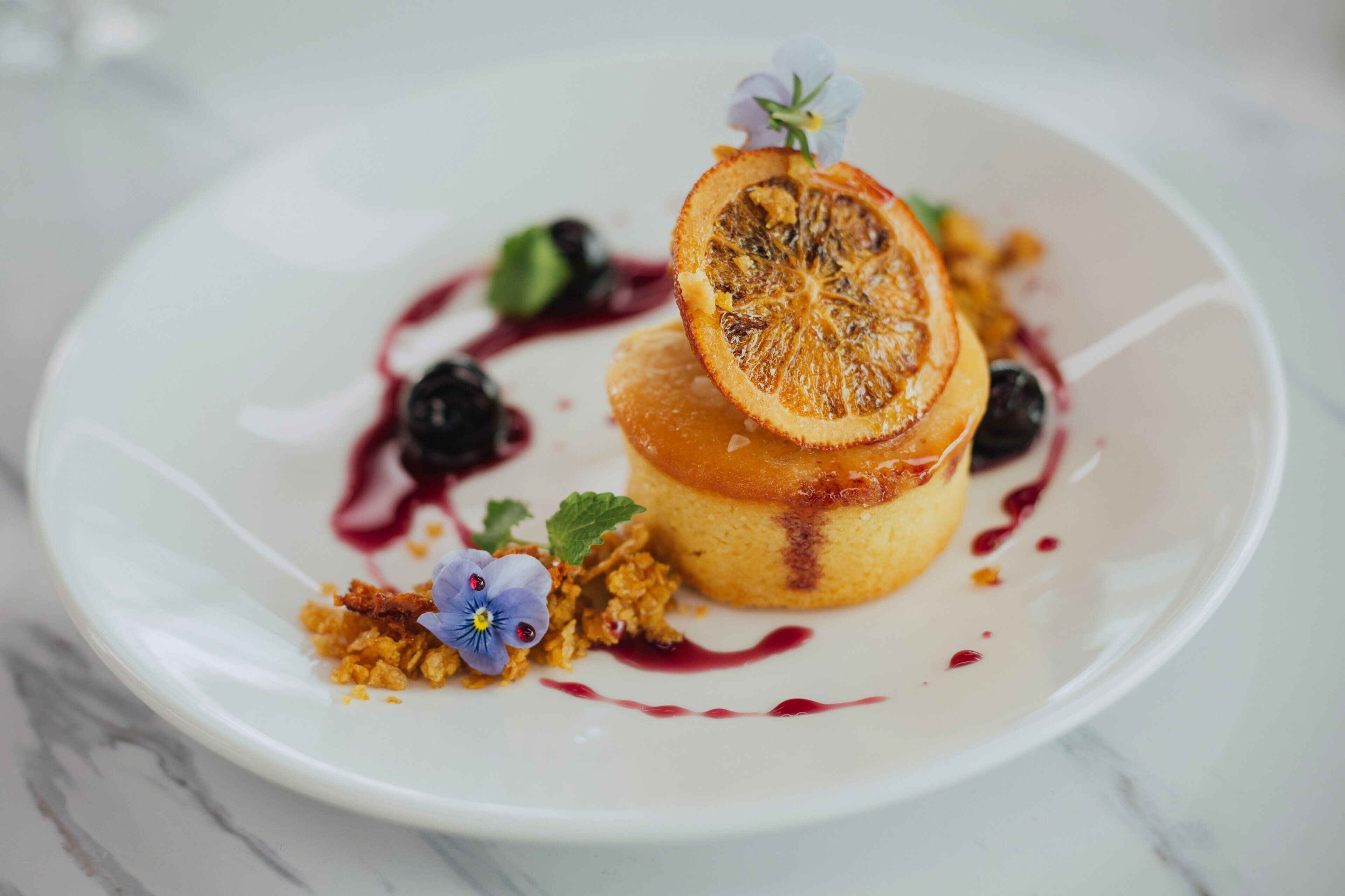 A dessert on a white plate with a slice of orange, blueberries, and a small flower.