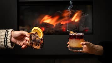 Dos manos sosteniendo una taza y un vaso con bebida, frente a una chimenea.