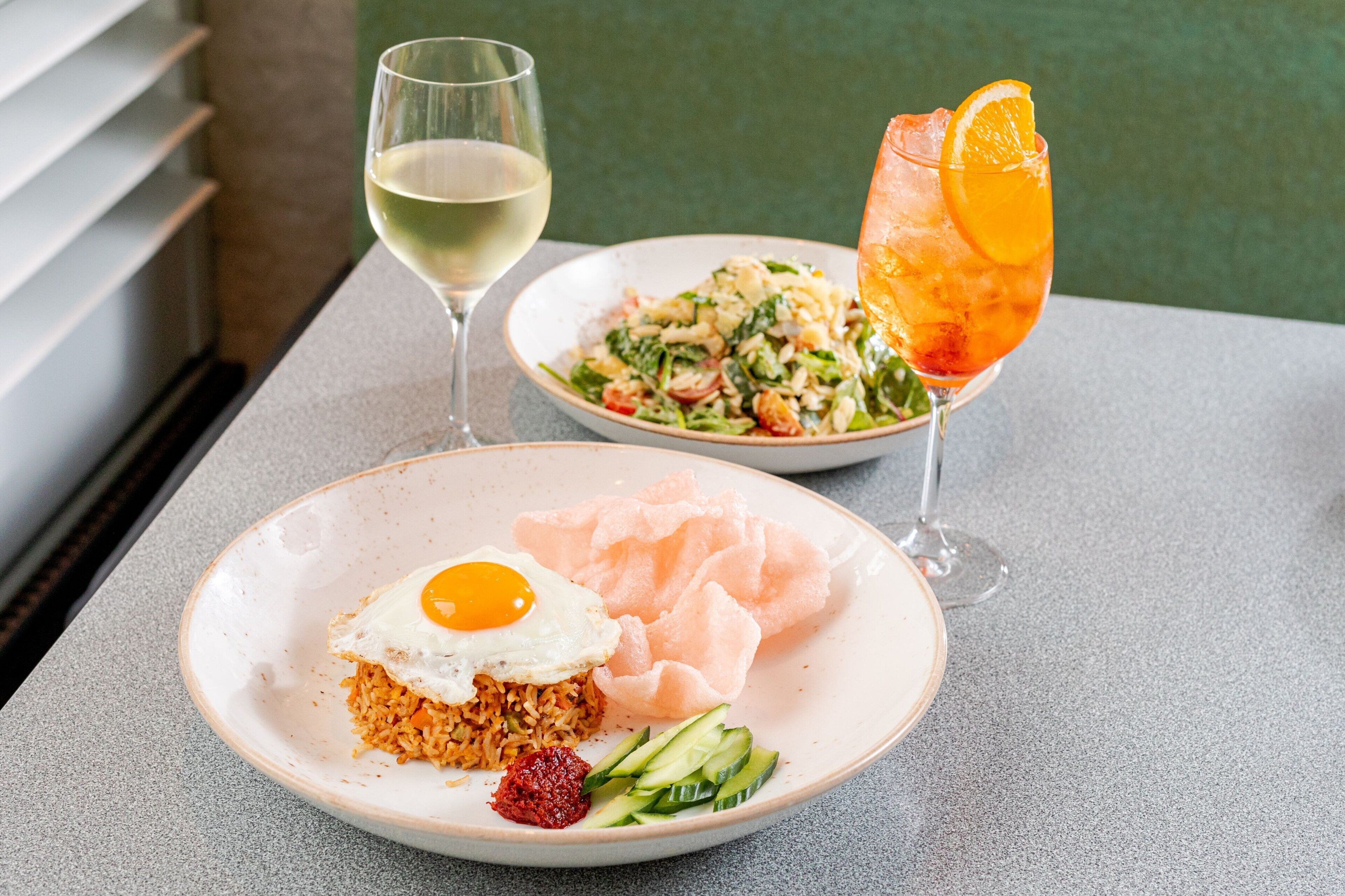 A table with a plate of food, a glass of white wine, and a cocktail with an orange slice.
