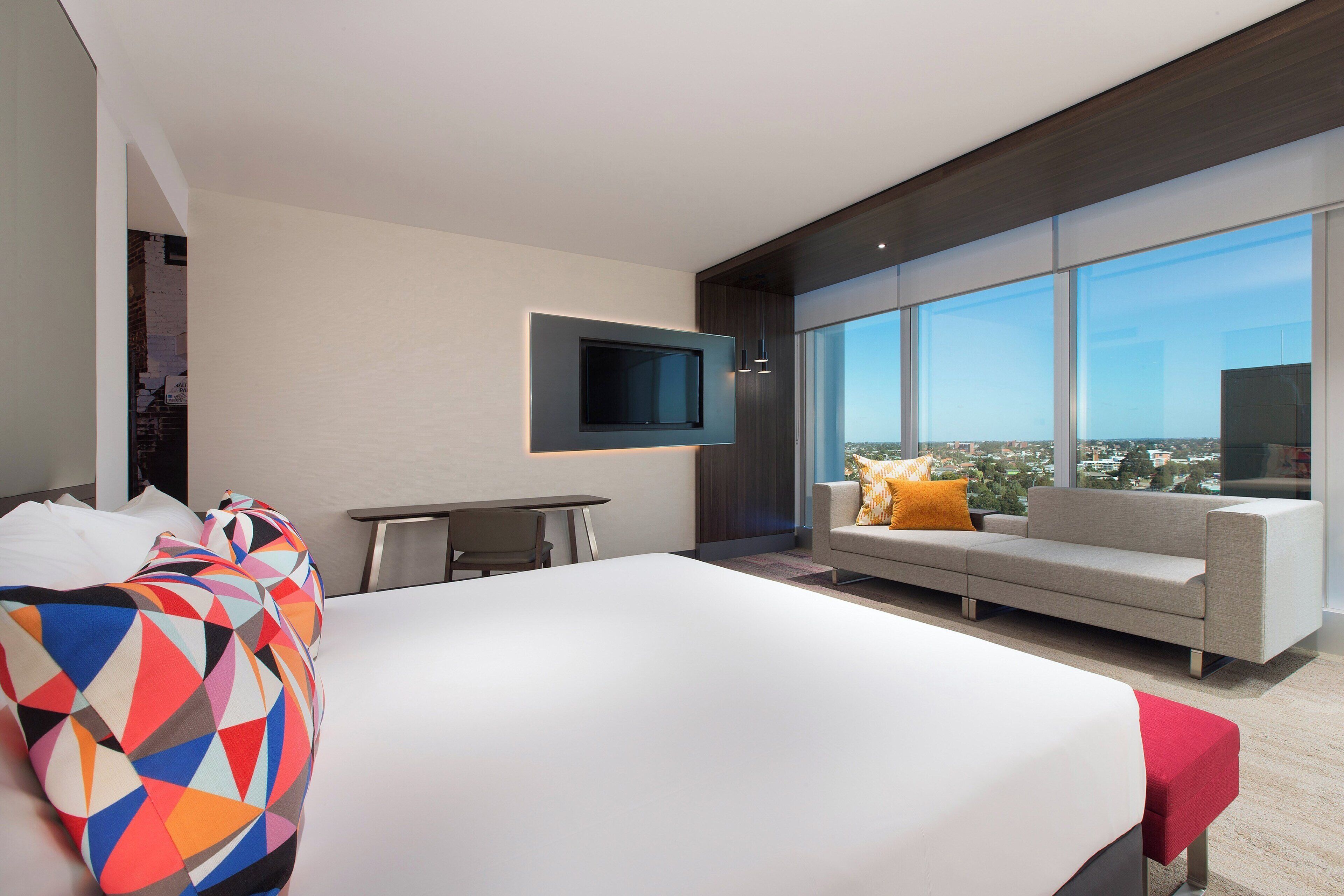 A modern hotel room with a large bed, a sofa, a TV mounted on the wall, and a city view through large windows.