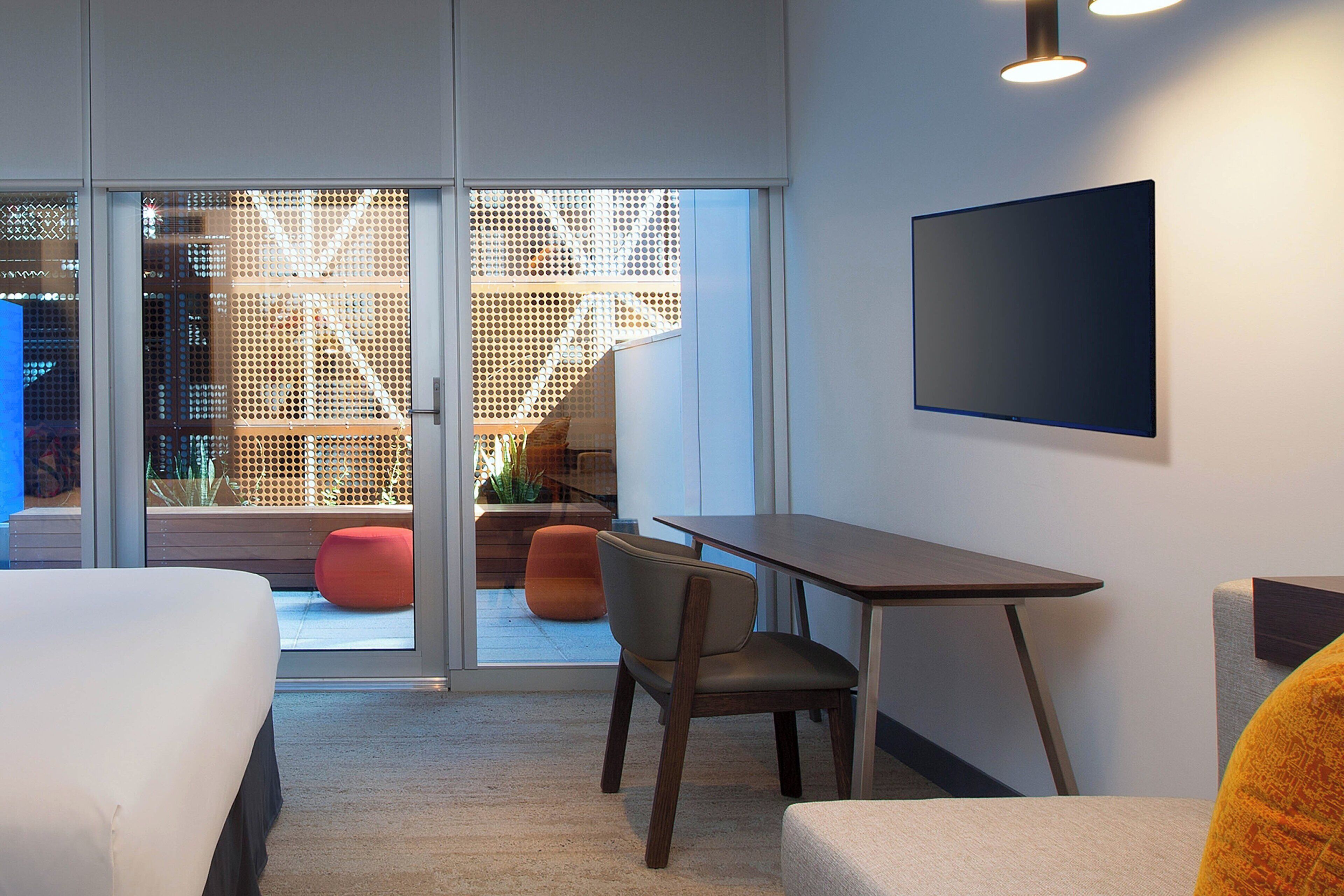 A modern hotel room with a bed, a desk, and a chair. There is a large window with a view of the outside.