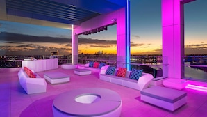 A modern rooftop lounge with white sofas and colorful cushions, large windows offering a city view at sunset. - Aloft by Marriott Perth (Rivervale)