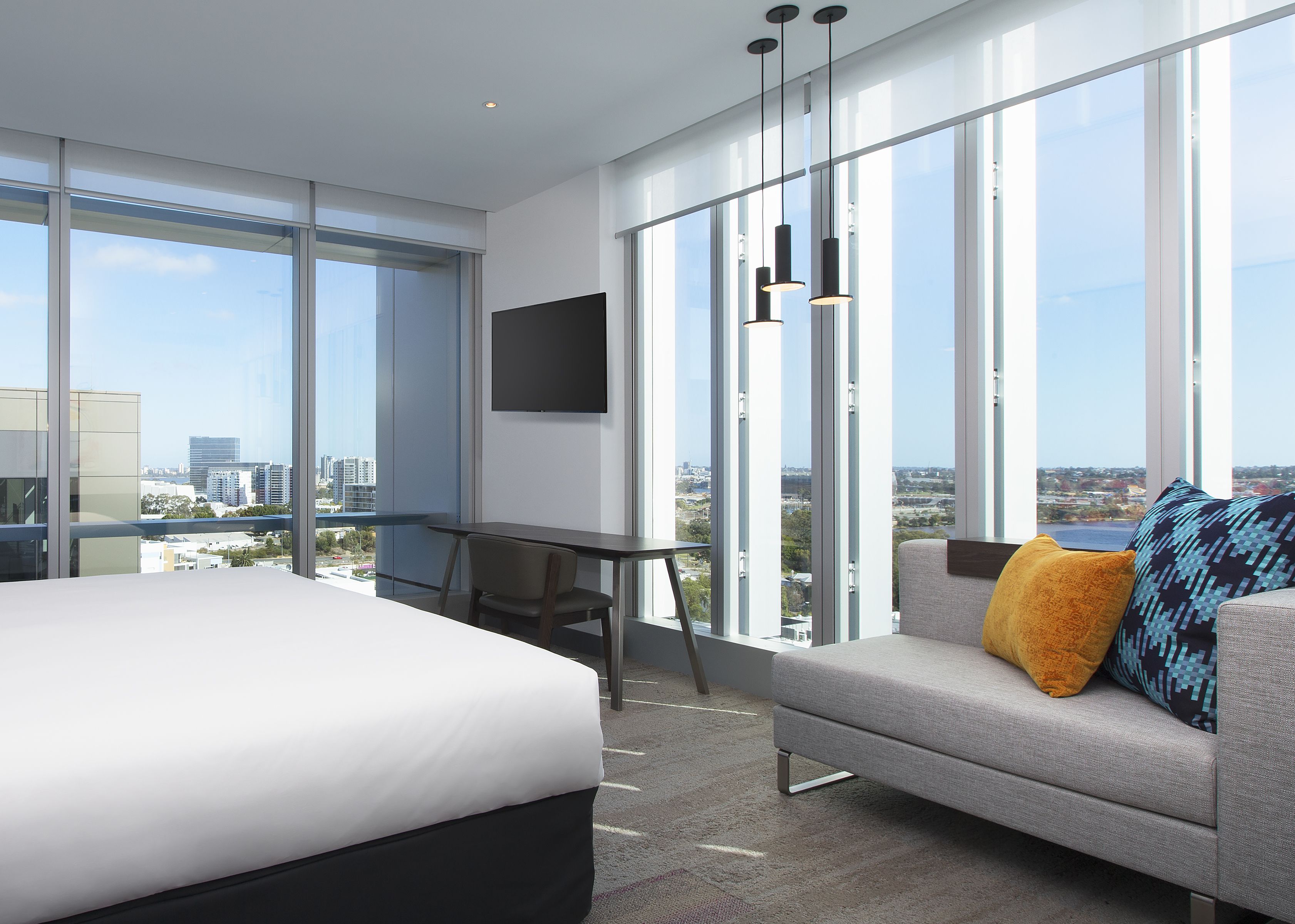 A modern hotel room with a large bed, a sofa with a patterned pillow, a desk, and a TV on the wall.