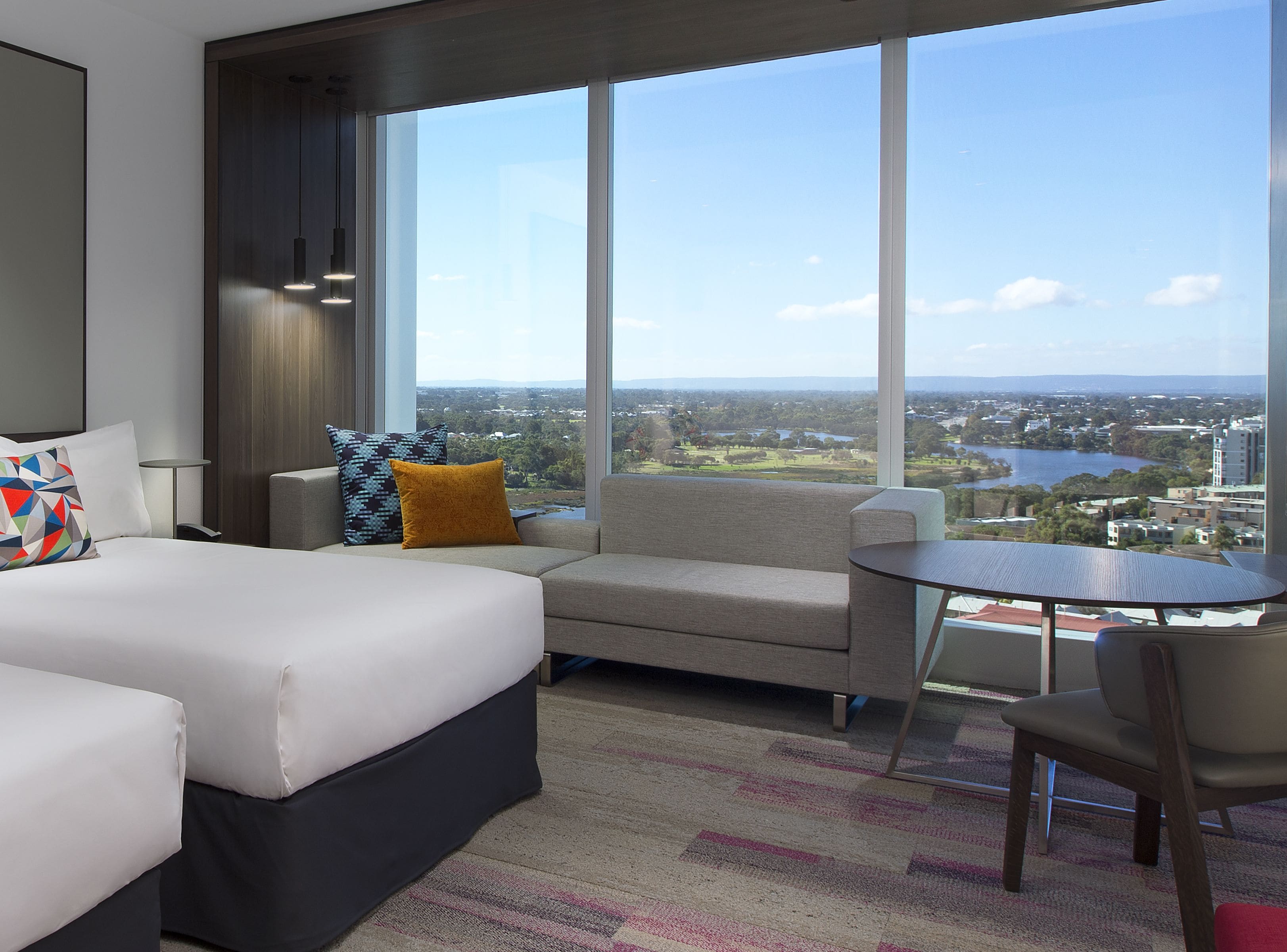 A hotel room with a bed, a sofa, a small table, and a chair. There is a large window offering a view of a cityscape and a body of water.