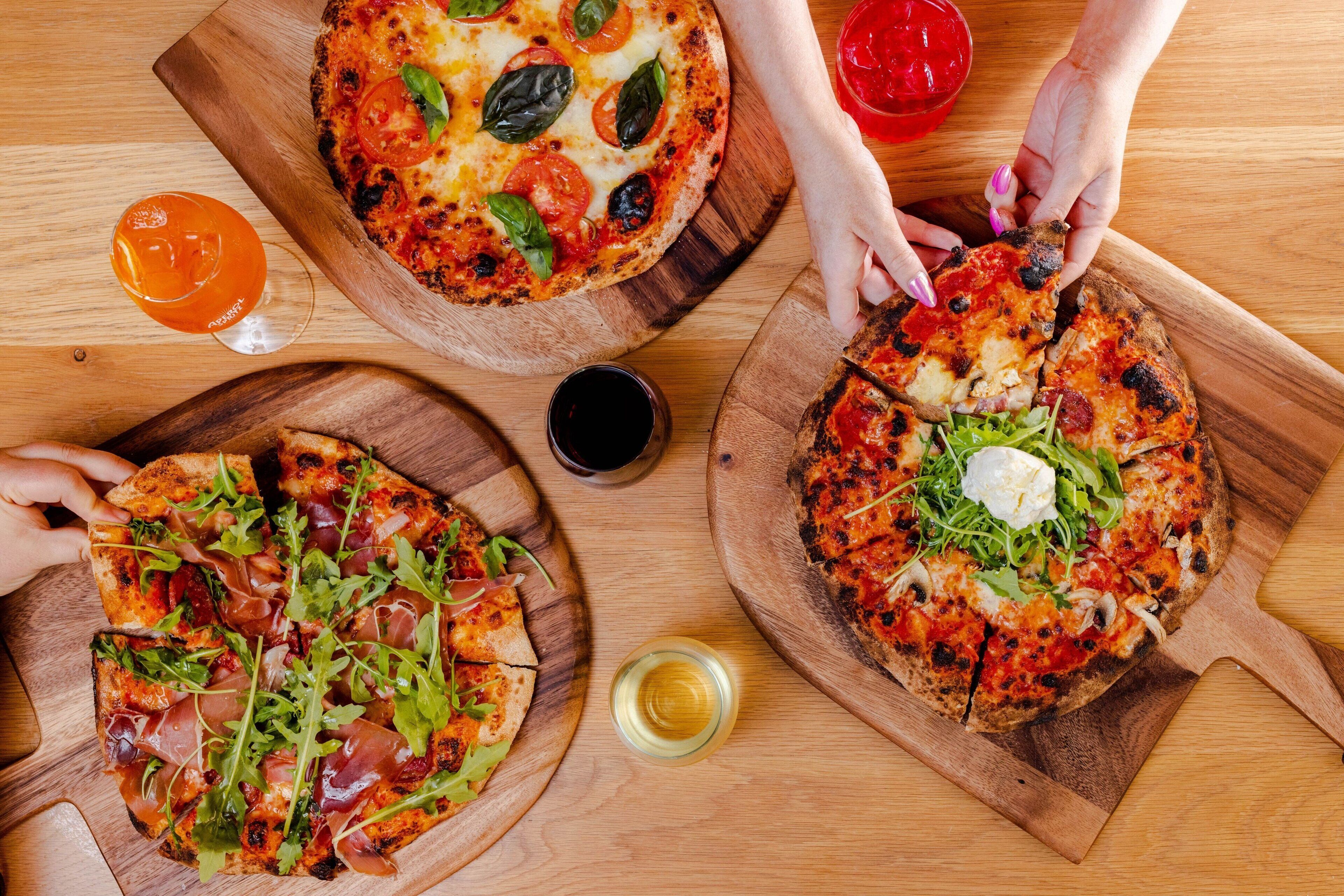 Tres pizzas sobre tablas de madera, con diferentes ingredientes y adornos, acompañadas de vasos de bebidas.