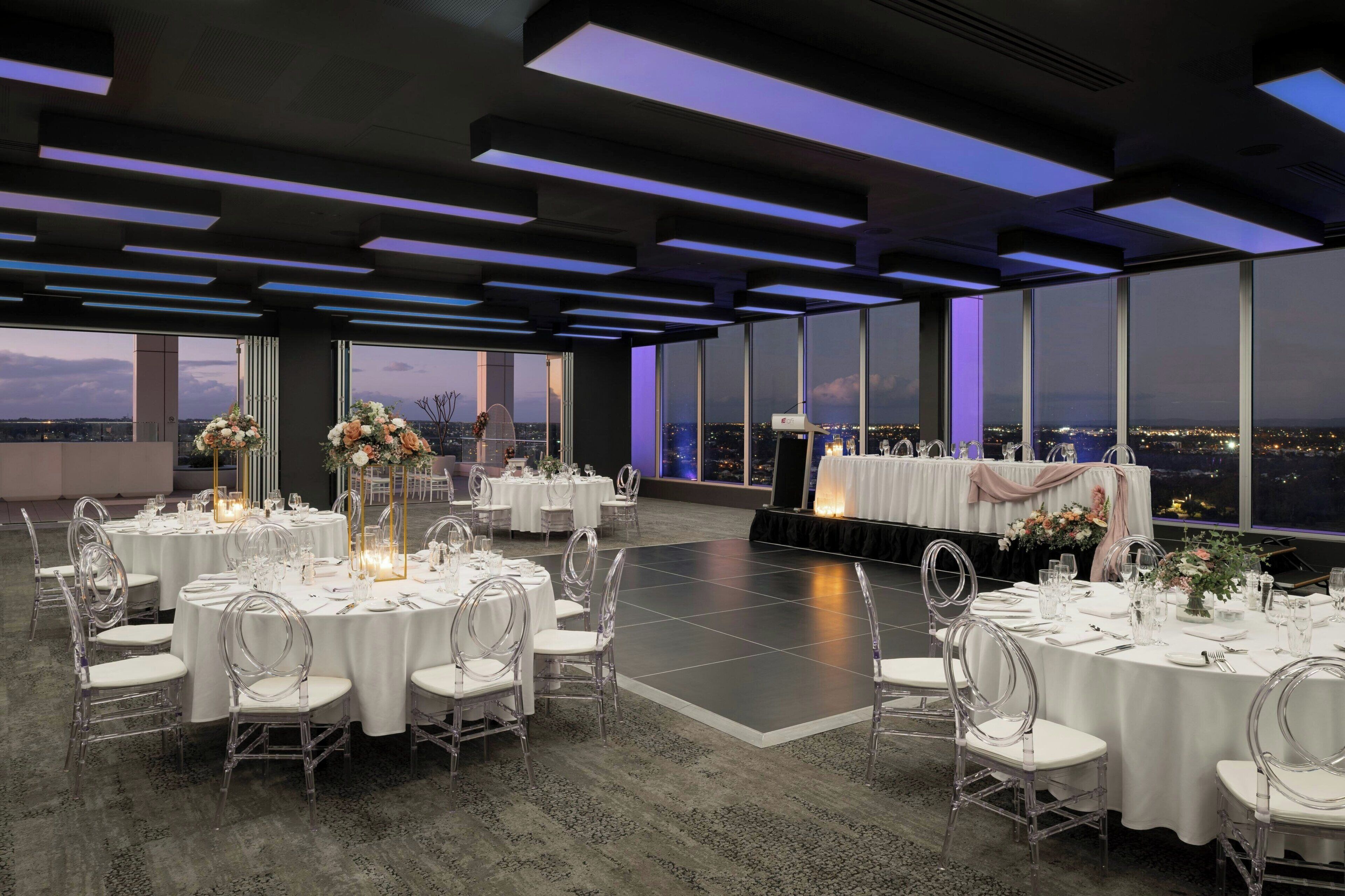Un espacio amplio para eventos con mesas redondas dispuestas para una ocasión formal, grandes ventanales con vistas a la ciudad y un escenario con un telón de fondo.
