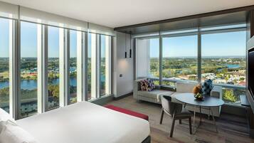 A modern hotel room with a large bed, a dining area, and a view of a lake and cityscape through floor-to-ceiling windows.
