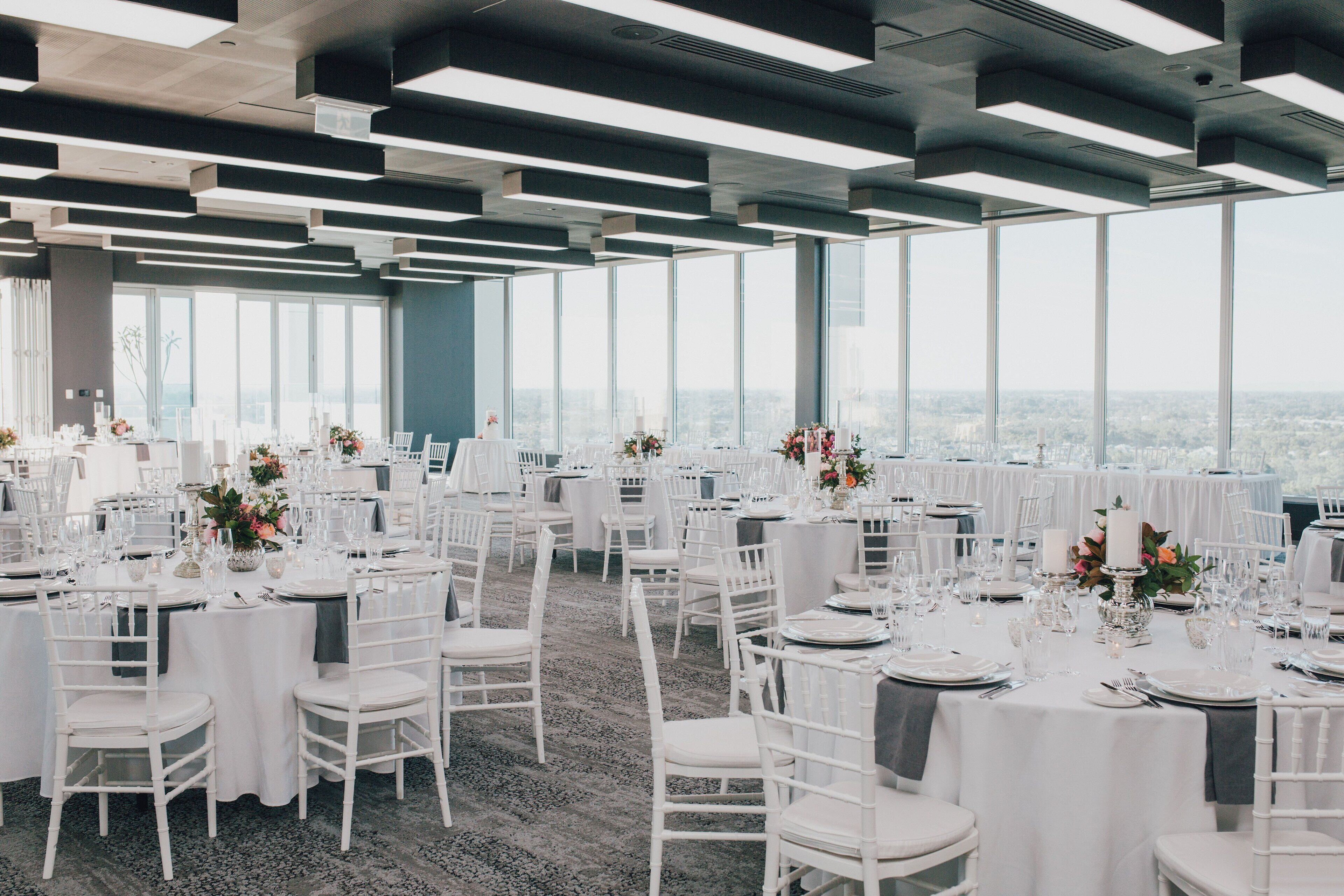 A spacious event space with multiple round tables set for dining, large windows offering a city view, and a modern ceiling design.