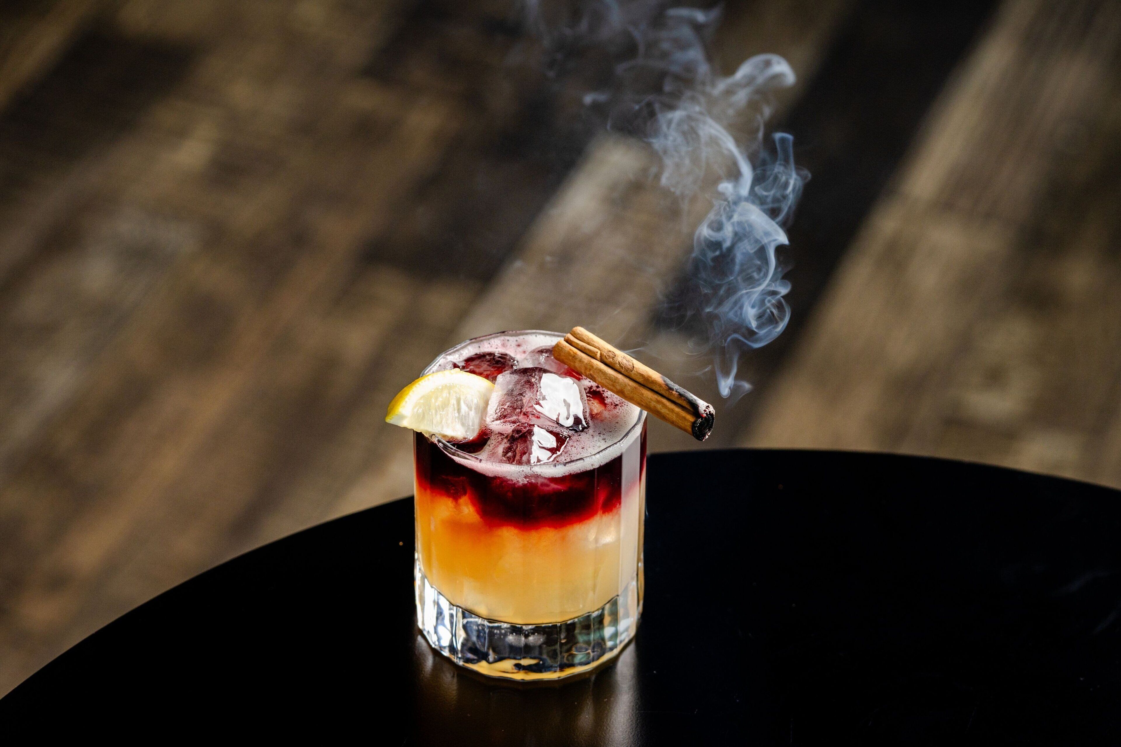 A cocktail with a lemon slice and a cinnamon stick on the rim, with smoke rising from the drink.