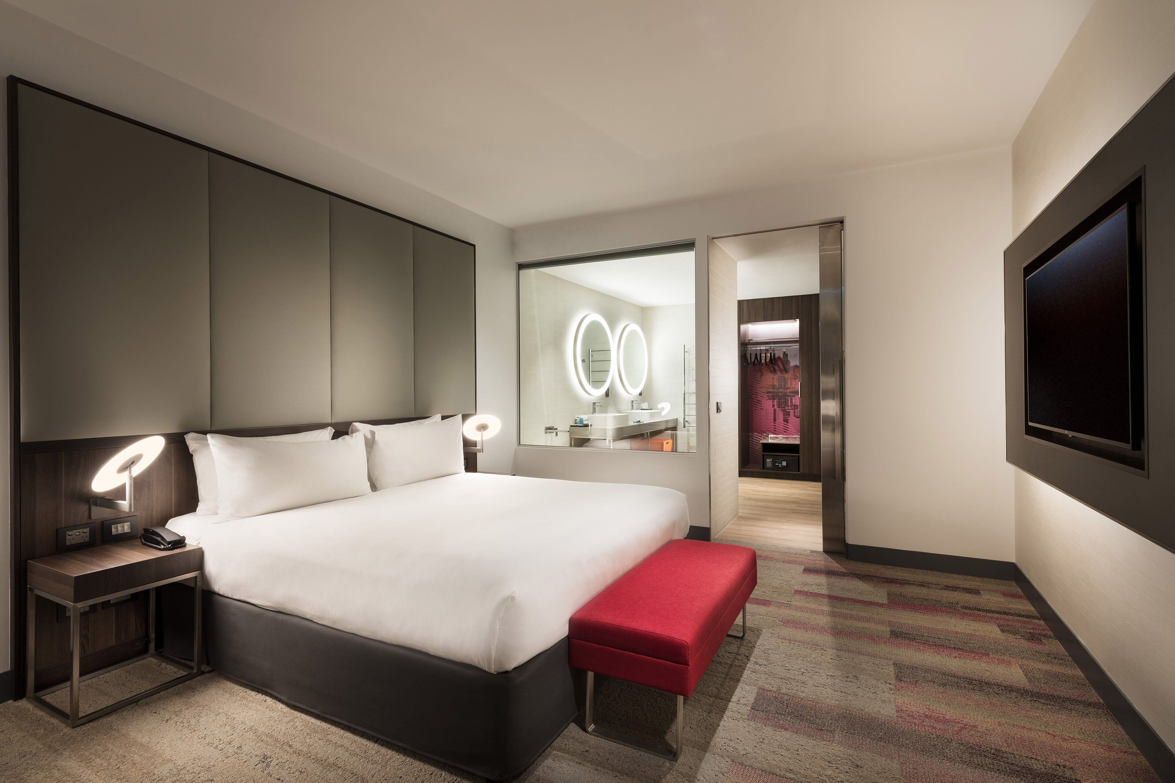 A modern hotel room with a large bed, bedside lamps, a bench, a TV, and a mirror with lights.