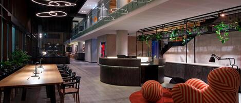 A spacious lobby with a long wooden reception desk, modern seating, and a staircase with greenery.