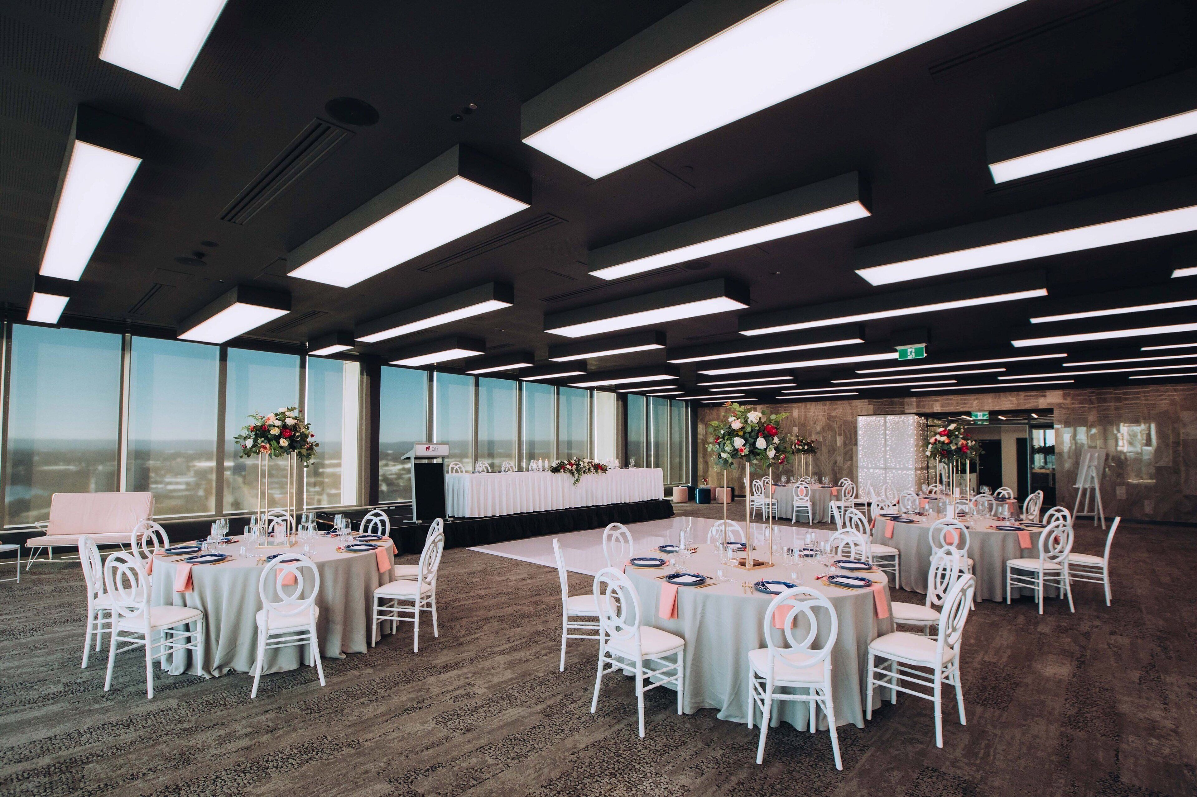 A spacious event room with multiple round tables, white chairs, and large windows offering a view.