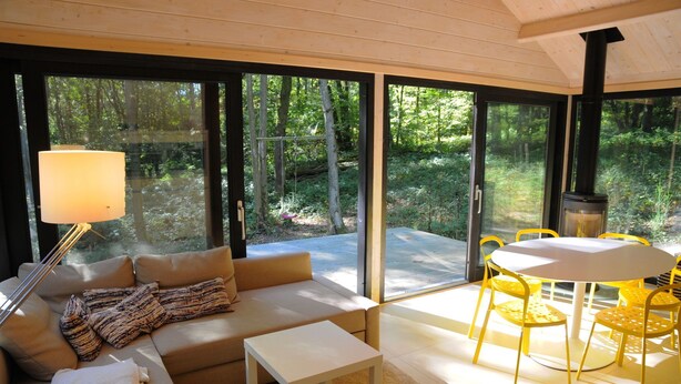 A modern living room with a sofa, coffee table, and yellow chairs. Large windows offer a view of the forest. - Chalet in Oignies near French Border (Oignies en Thierache)