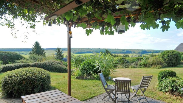 Eine Holzdielen-Terrasse mit Tisch und zwei Stühlen, mit Blick auf einen üppigen Garten und die entfernten Hügel.