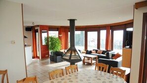 Ein Essbereich mit einem runden Tisch, Stühlen und einem Kamin im Zentrum. Im Hintergrund befinden sich eine Couch und ein Fernseher.