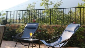Deux fauteuils de jardin avec un verre posé sur une table, sur une terrasse dotée d’un mur en pierre et d’une clôture en métal.
