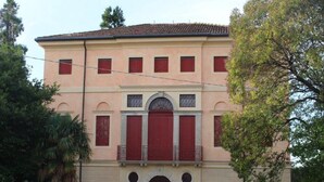 Un bâtiment à deux étages doté d’une façade symétrique, avec une entrée centrale surmontée d’un arc décoratif, flanquée de deux colonnes, et une série de fenêtres avec des volets rouges.
