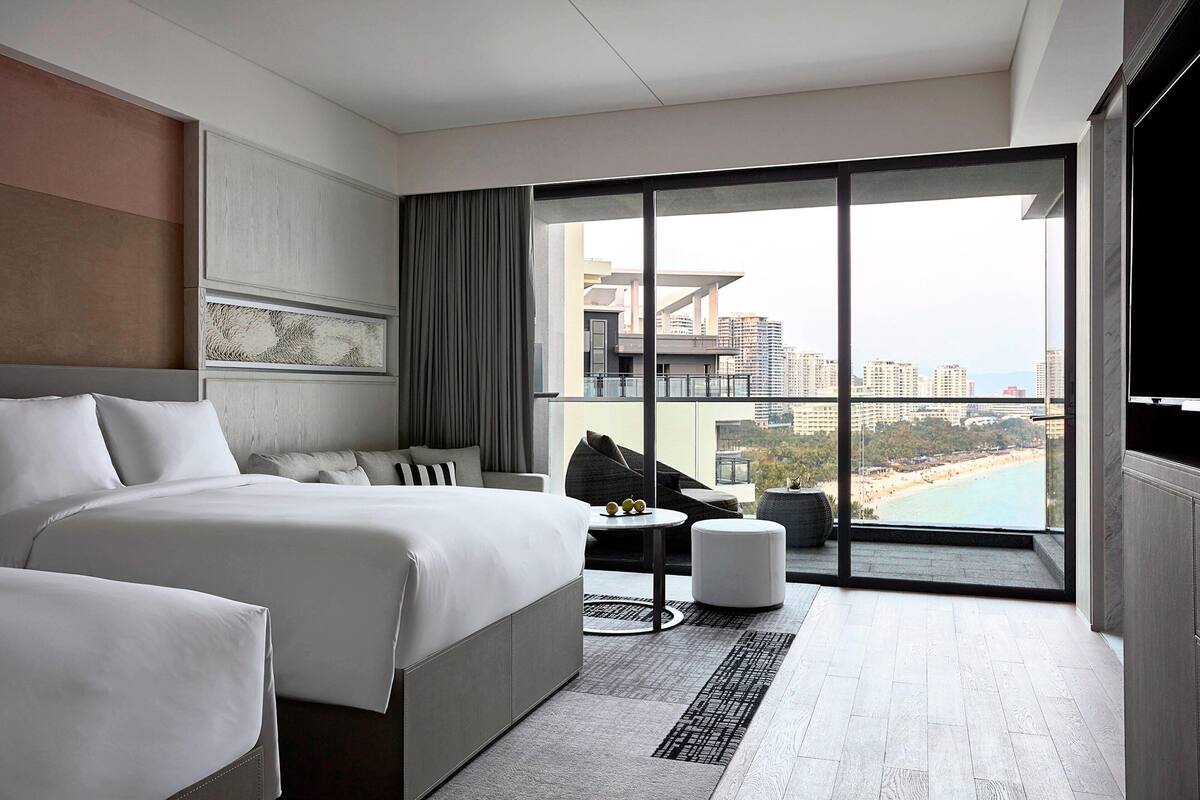 Une chambre d’hôtel moderne dotée d’un grand lit, d’un canapé, d’une petite table et offrant une vue sur la plage et le paysage urbain grâce à de grandes fenêtres du sol au plafond.