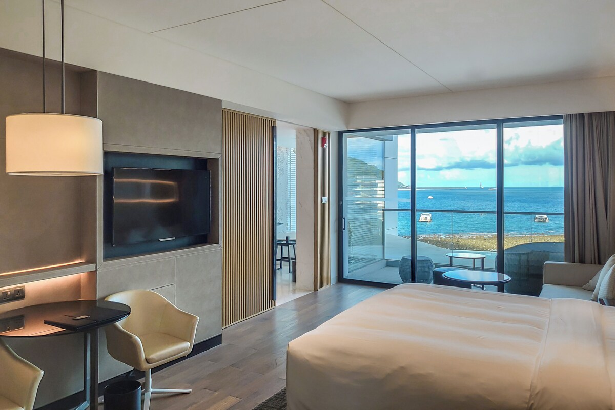 A modern hotel room with a large bed, a TV, a desk, and a balcony with a view of the sea.