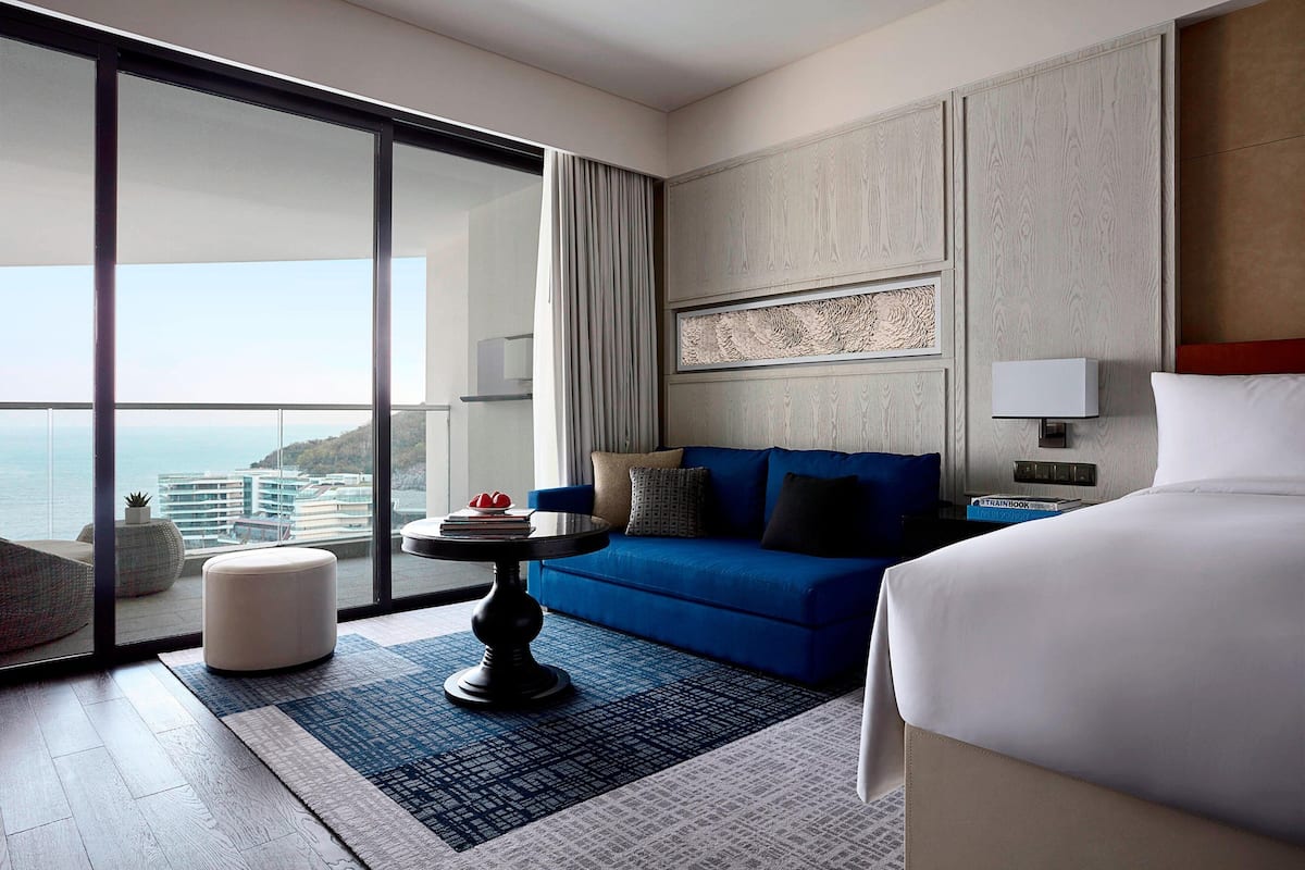 Une chambre d’hôtel moderne avec un grand lit, un canapé bleu, une petite table ronde et une vue sur la ville grâce à de grandes fenêtres du sol au plafond.