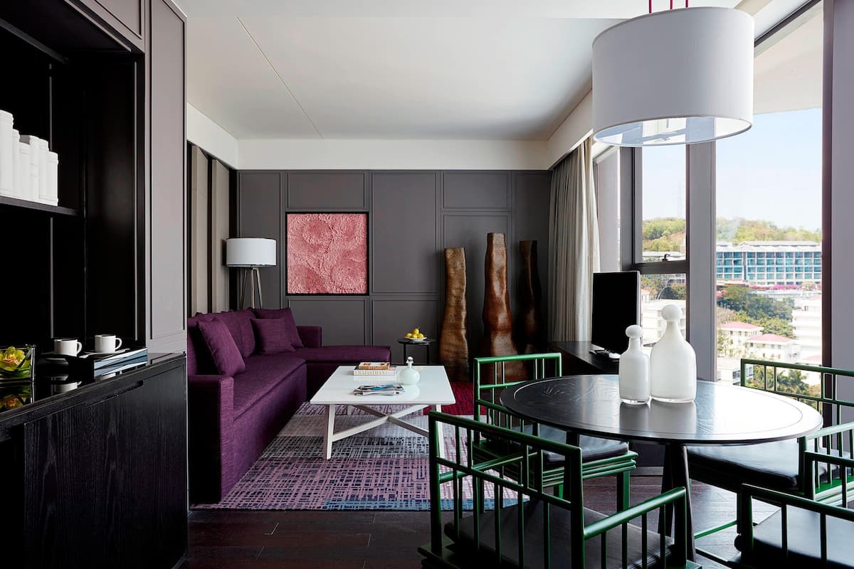 Une chambre d’hôtel moderne avec un canapé violet, une table basse blanche et un coin repas comprenant une table ronde et des chaises.