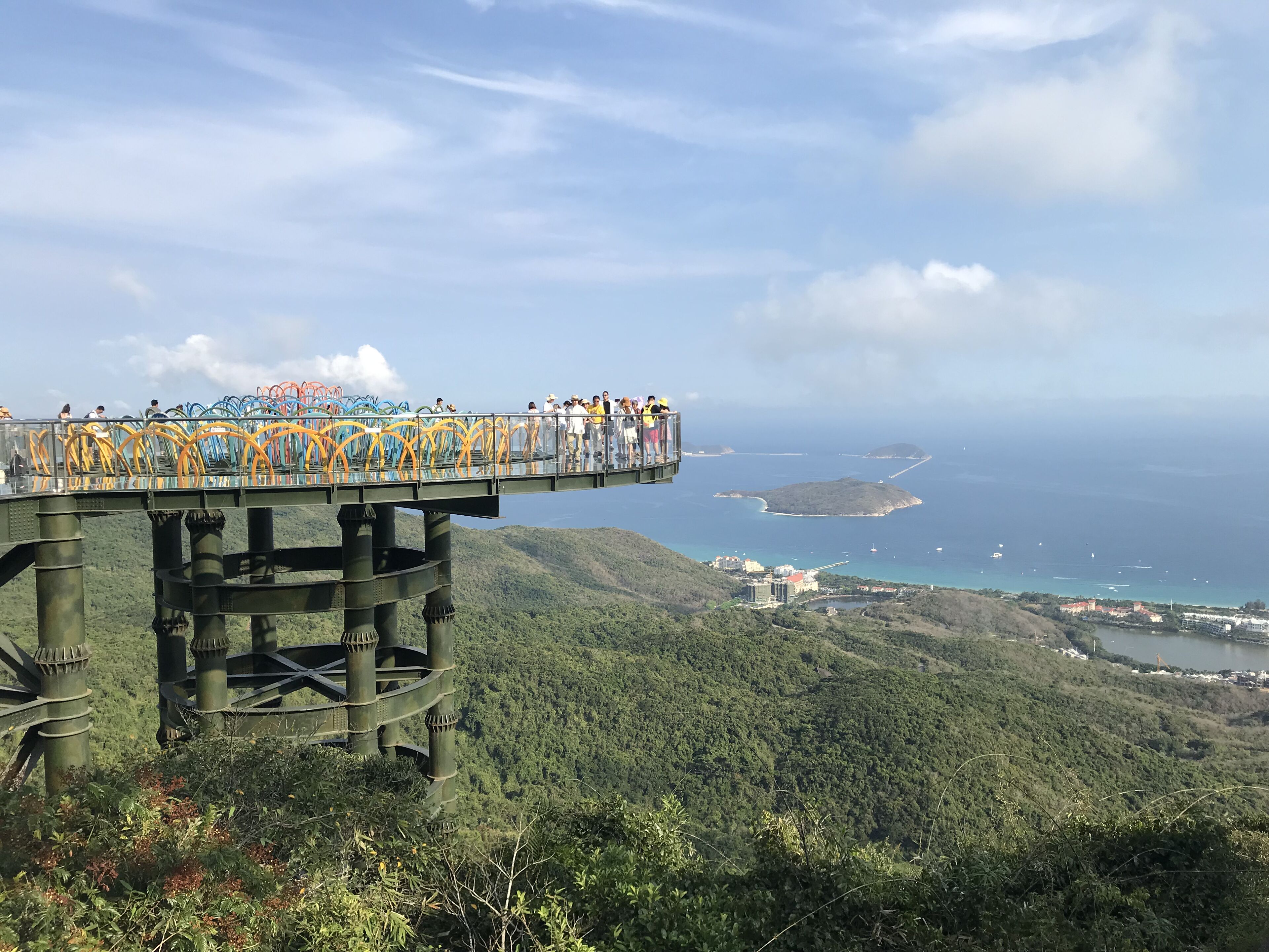 A glass bridge with people walking over it, overlooking a coastal landscape with hills and a body of water.