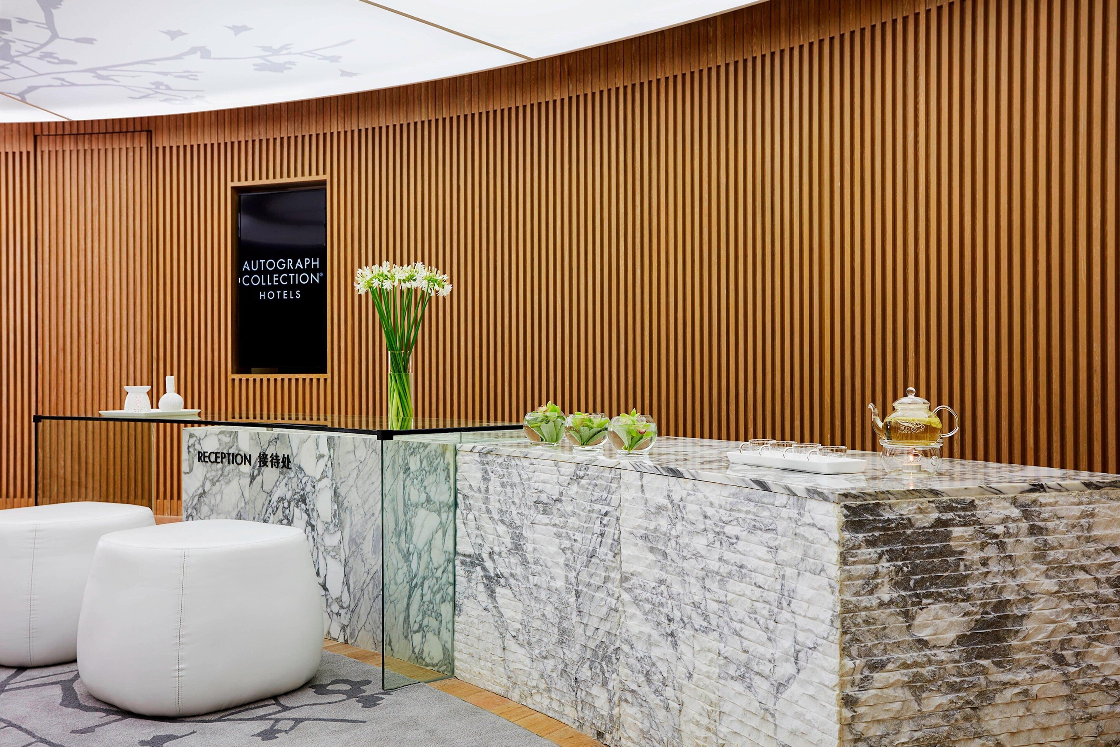 A reception desk with a marble top, a wooden panel wall, and a reception sign. There are three white cushioned seating areas and a vase with white flowers on the desk.
