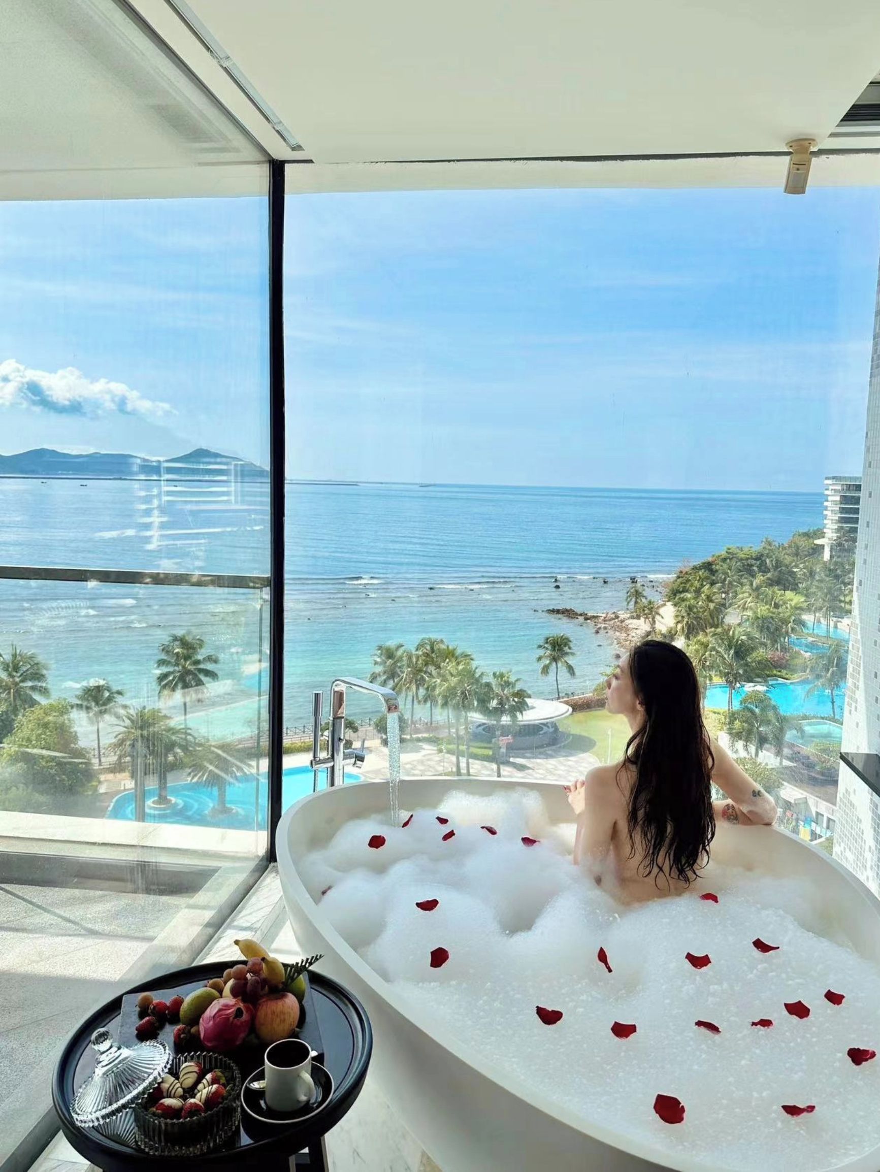 A person relaxing in a bathtub filled with bubbles and rose petals, overlooking a pool and ocean view.