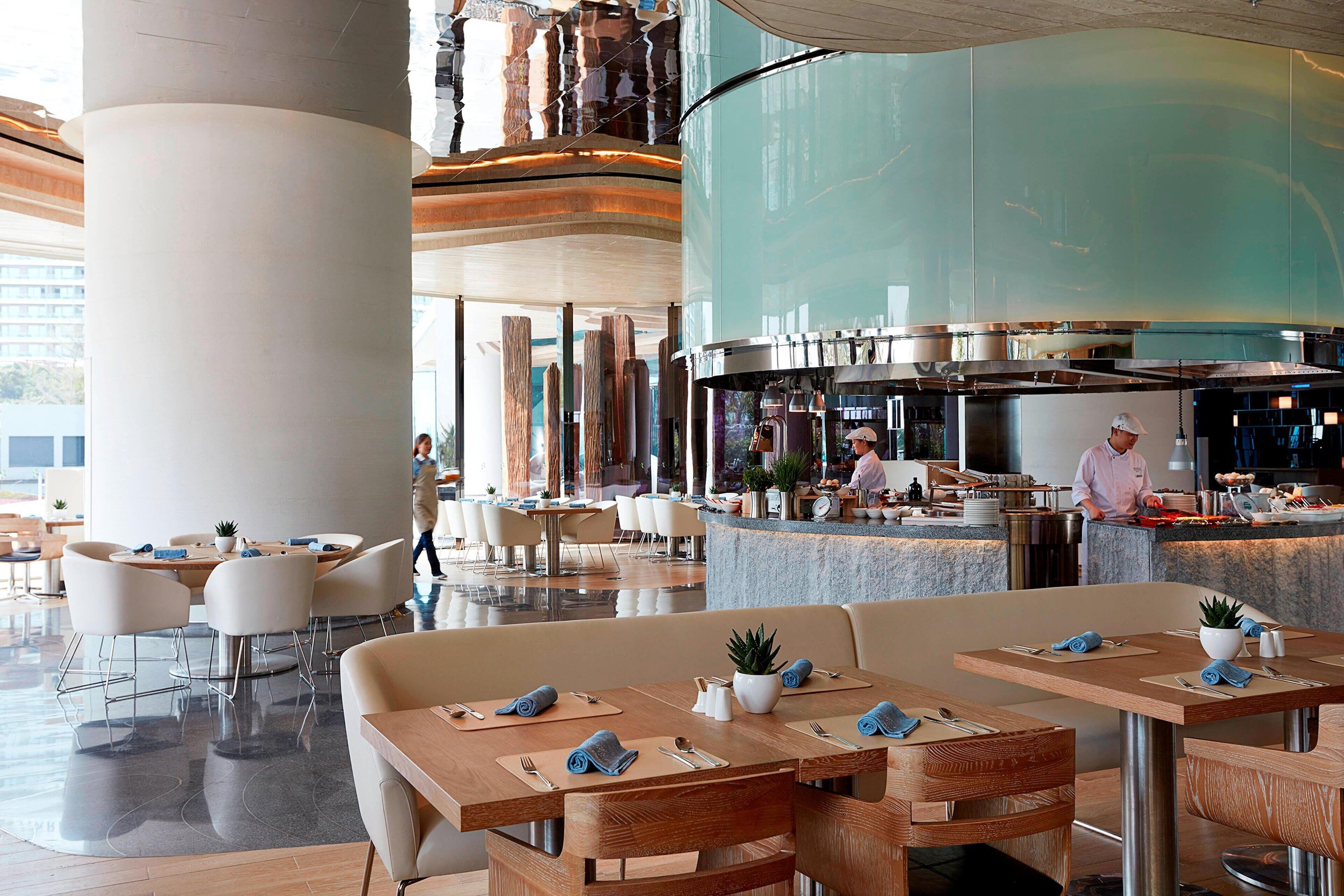 A modern hotel lobby with a central bar area, seating arrangements, and a visible kitchen in the background.