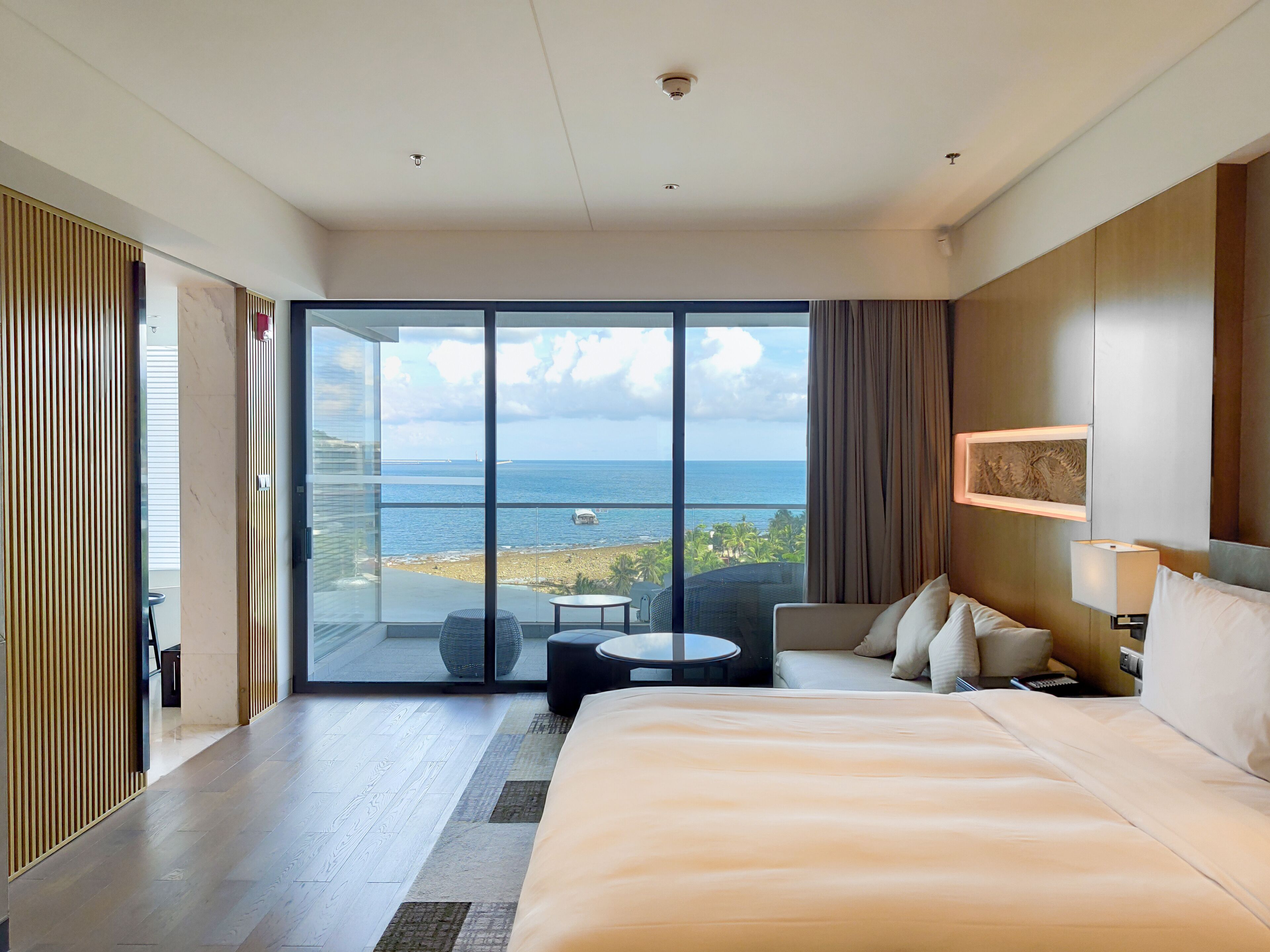 Habitación de hotel con una cama grande, una mesita pequeña, un sofá y vistas al océano a través de puertas correderas de vidrio.