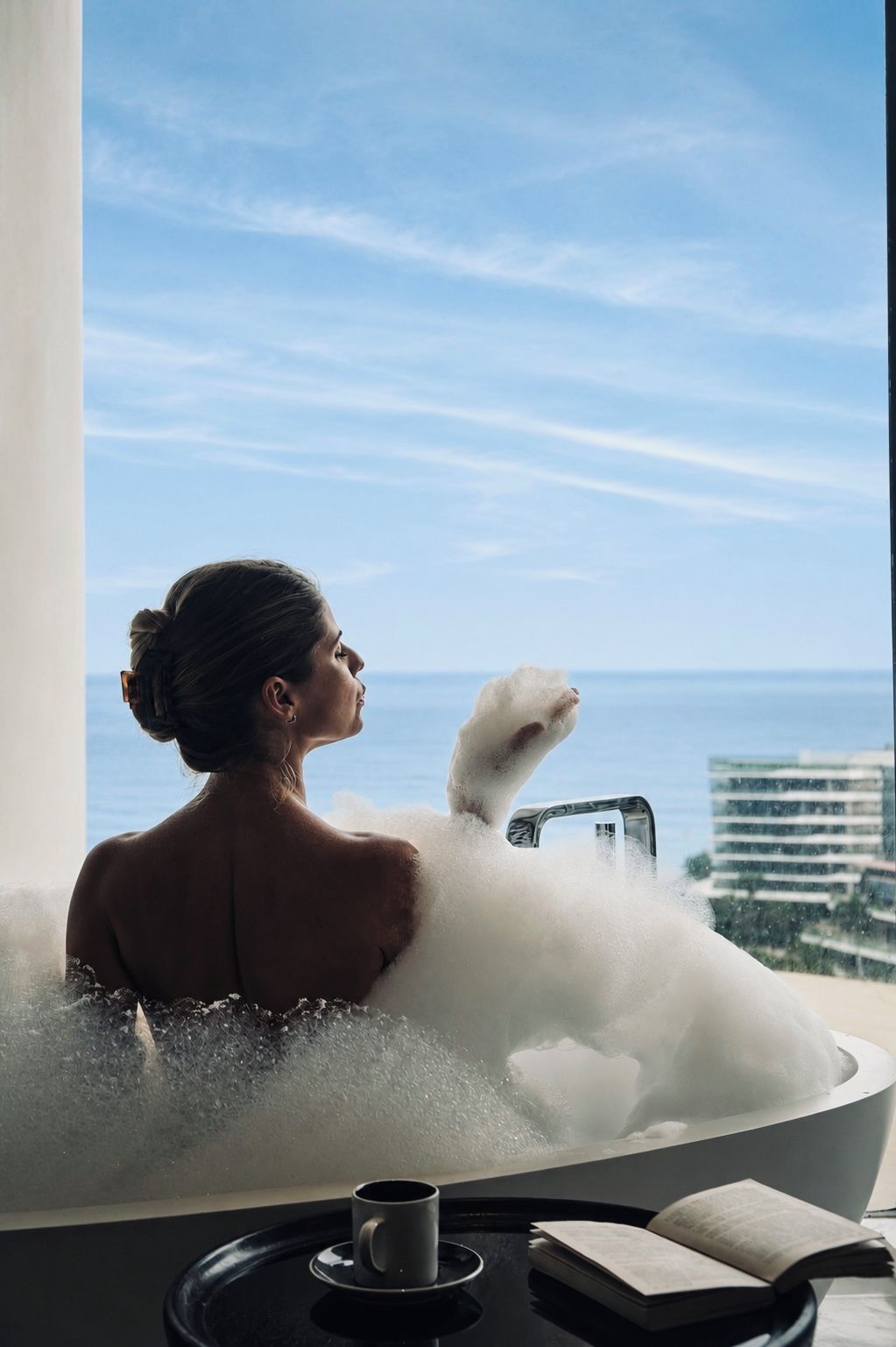 Eine Person liegt in einer mit Blasen gefüllten Badewanne, im Hintergrund Meer und Gebäude.