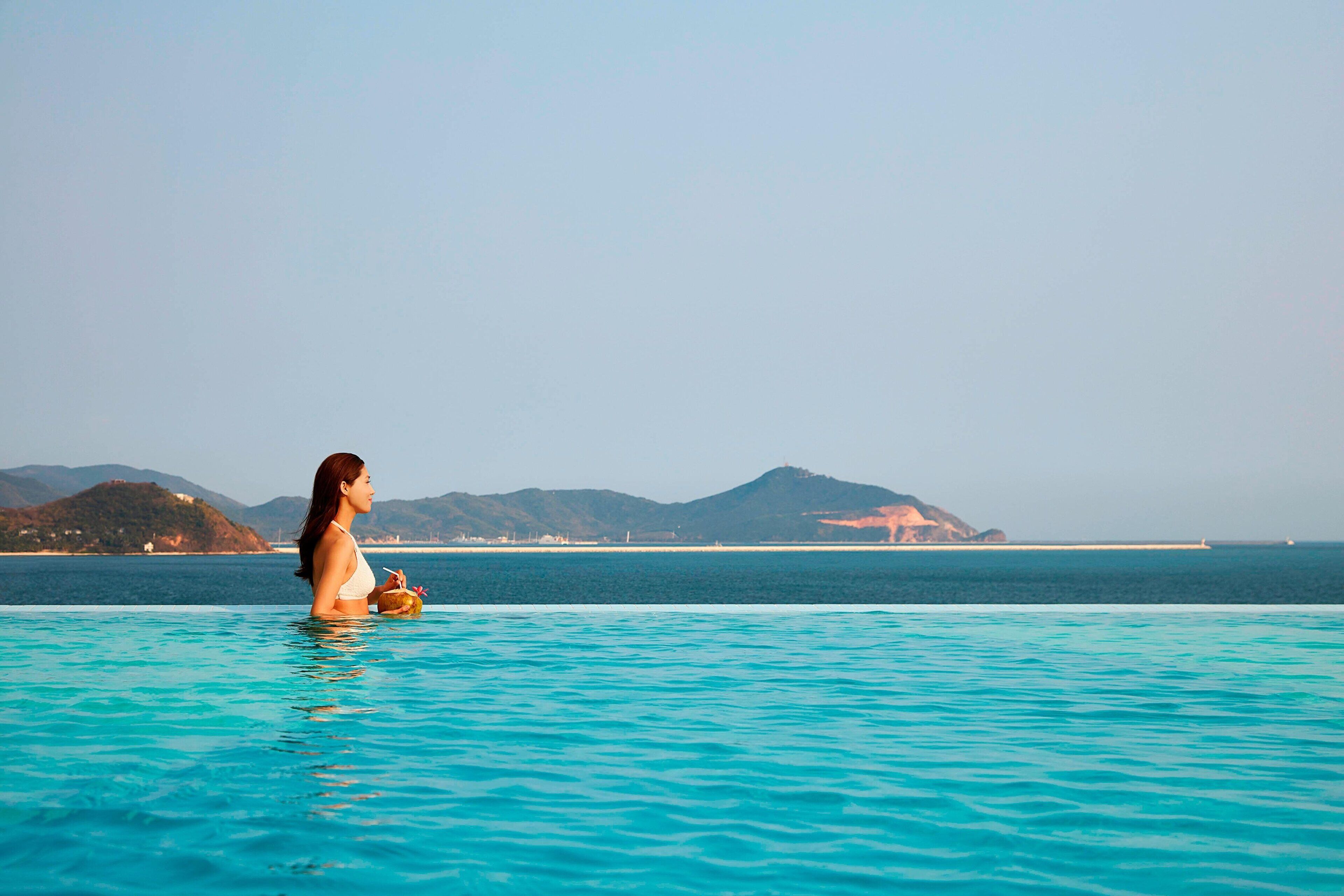 A woman in a swimming pool with a view of a distant coastline and mountains.