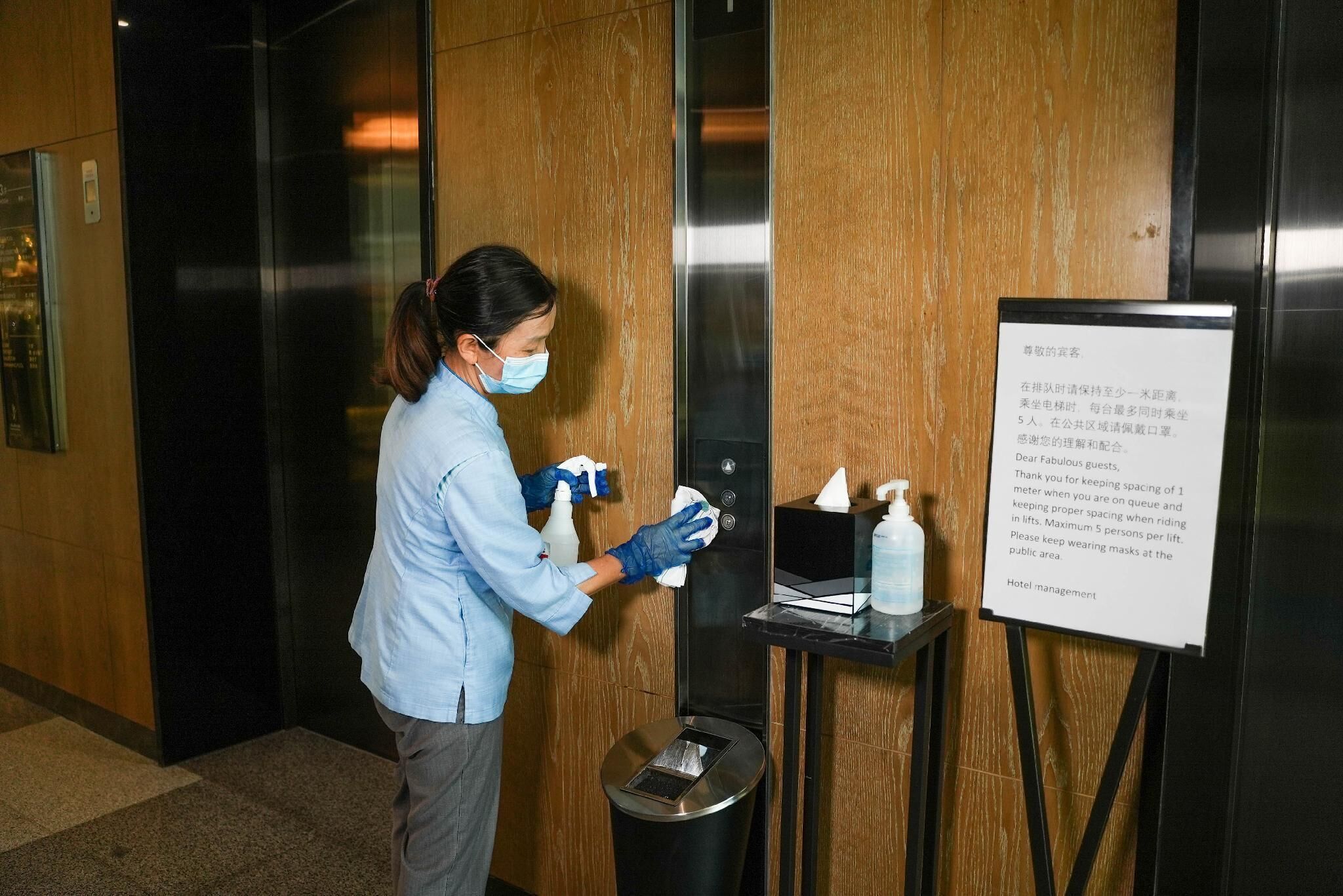 Una persona limpiando un ascensor con aerosol desinfectante y toallitas, un cartel con instrucciones y un cesto de basura.