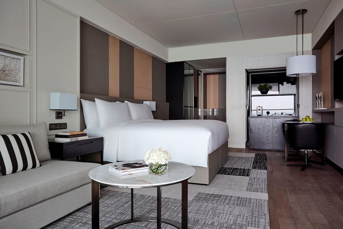 Une chambre d’hôtel moderne dotée d’un grand lit, d’un coin salon avec un canapé, d’une petite table ronde agrémentée d’un vase de fleurs, d’un bureau et d’un coin cuisine.