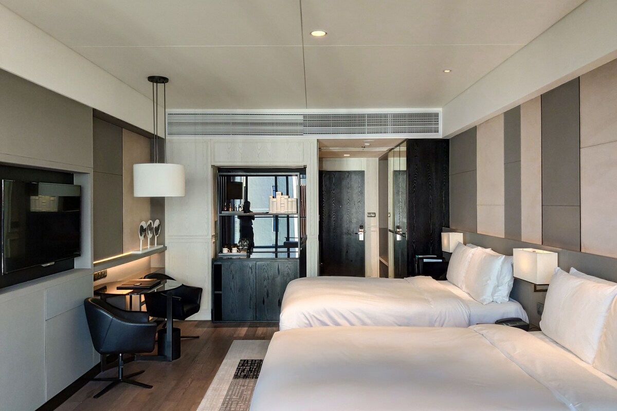 A modern hotel room with a large bed, a desk with a chair, a TV, and a bathroom visible through a glass door.