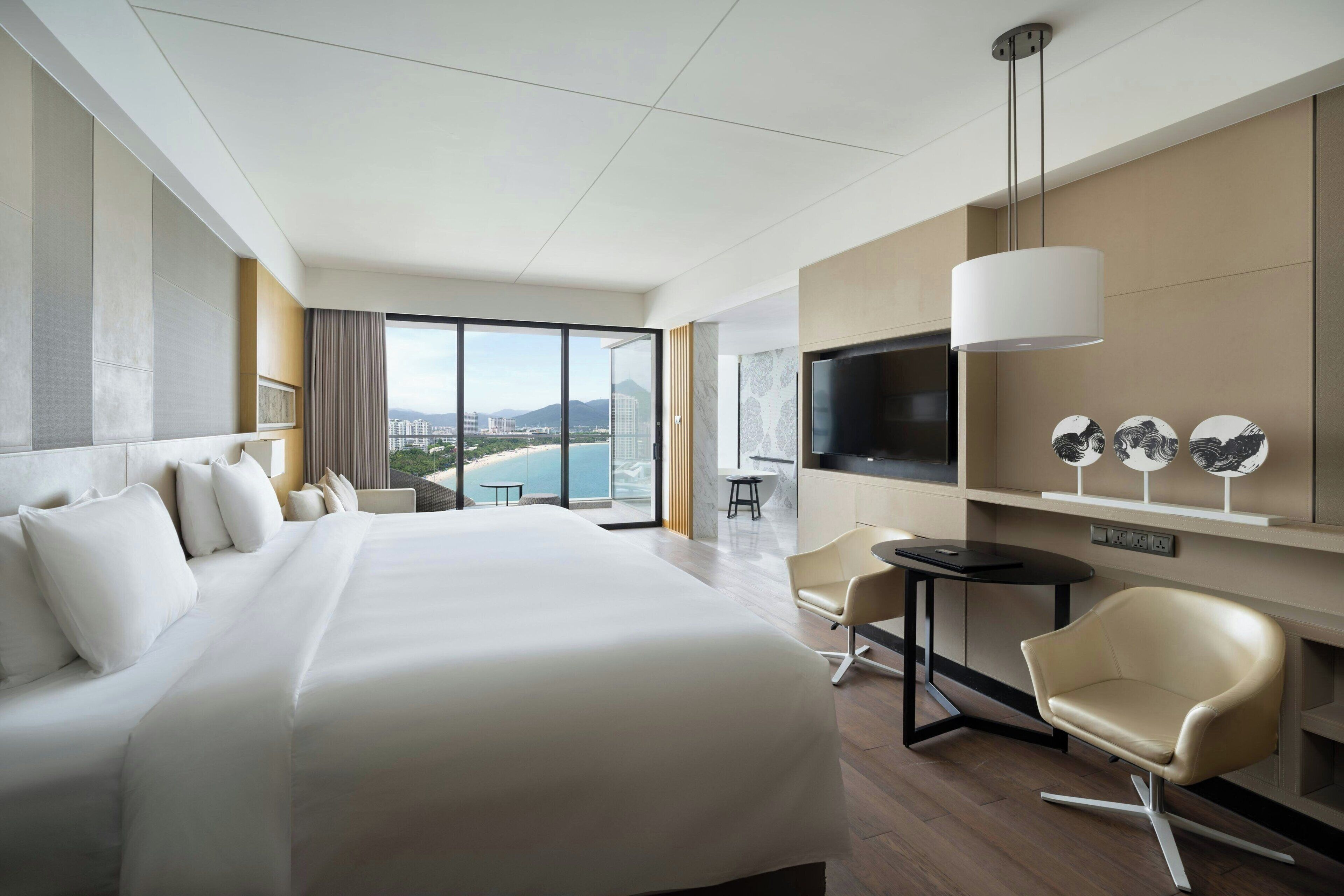 A modern hotel room with a large bed, a TV, a desk, and a chair. There is a view of the city and mountains through the windows.