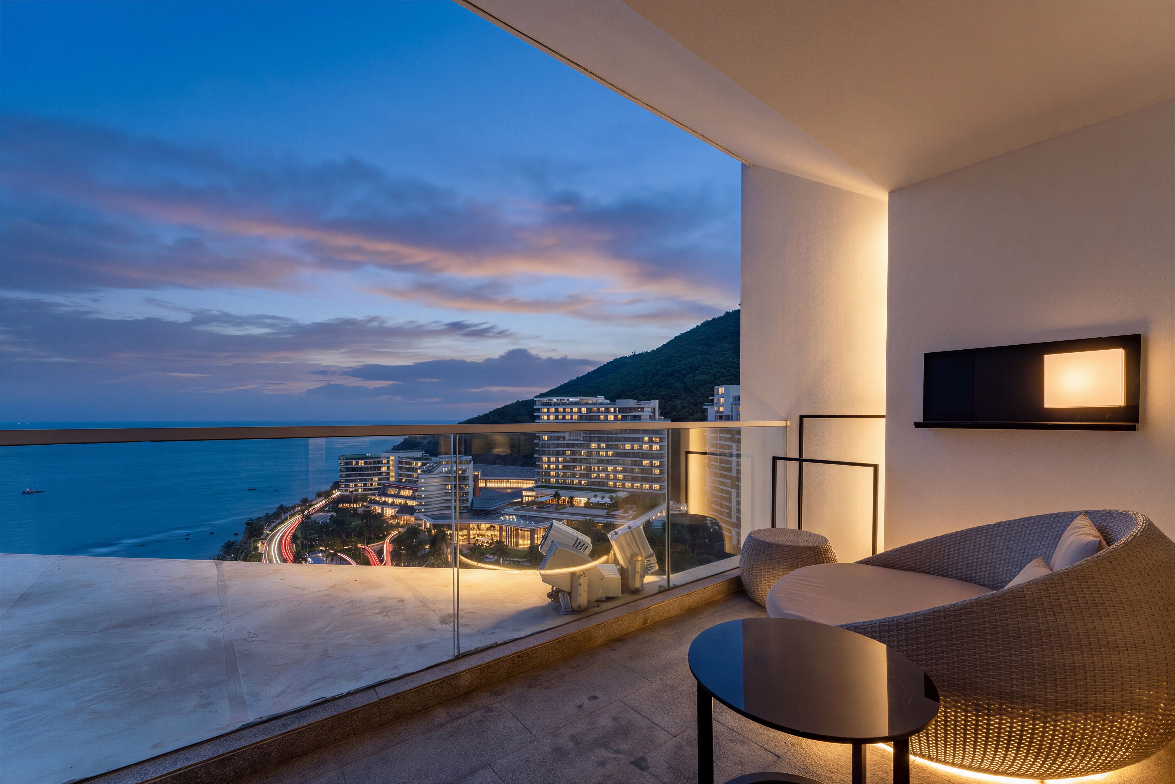 A balcony with a view of the sea and buildings, a sofa, a table, and a TV mounted on the wall.
