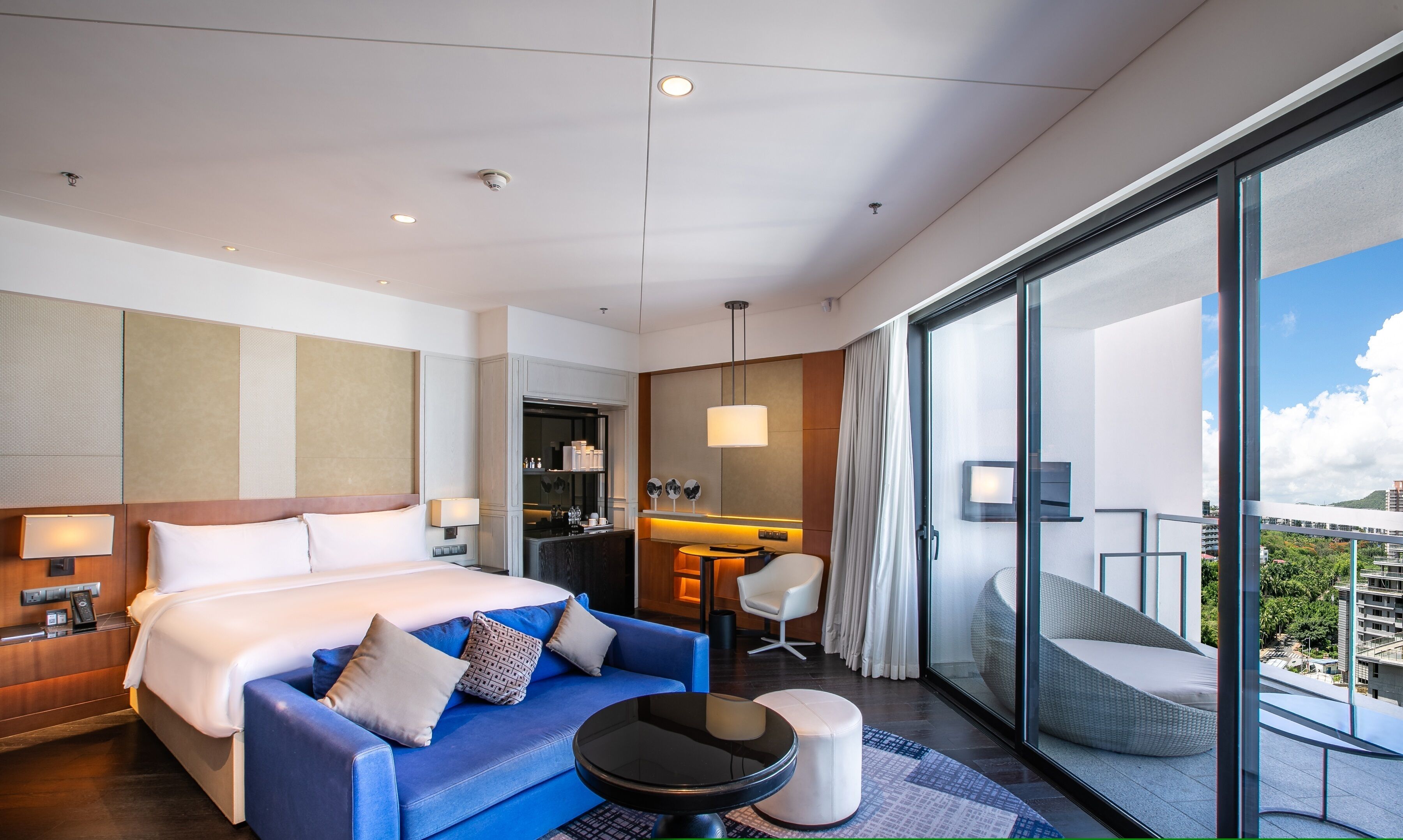 A modern hotel room with a large bed, a blue sofa, a small round table, and a balcony with a view of the city.