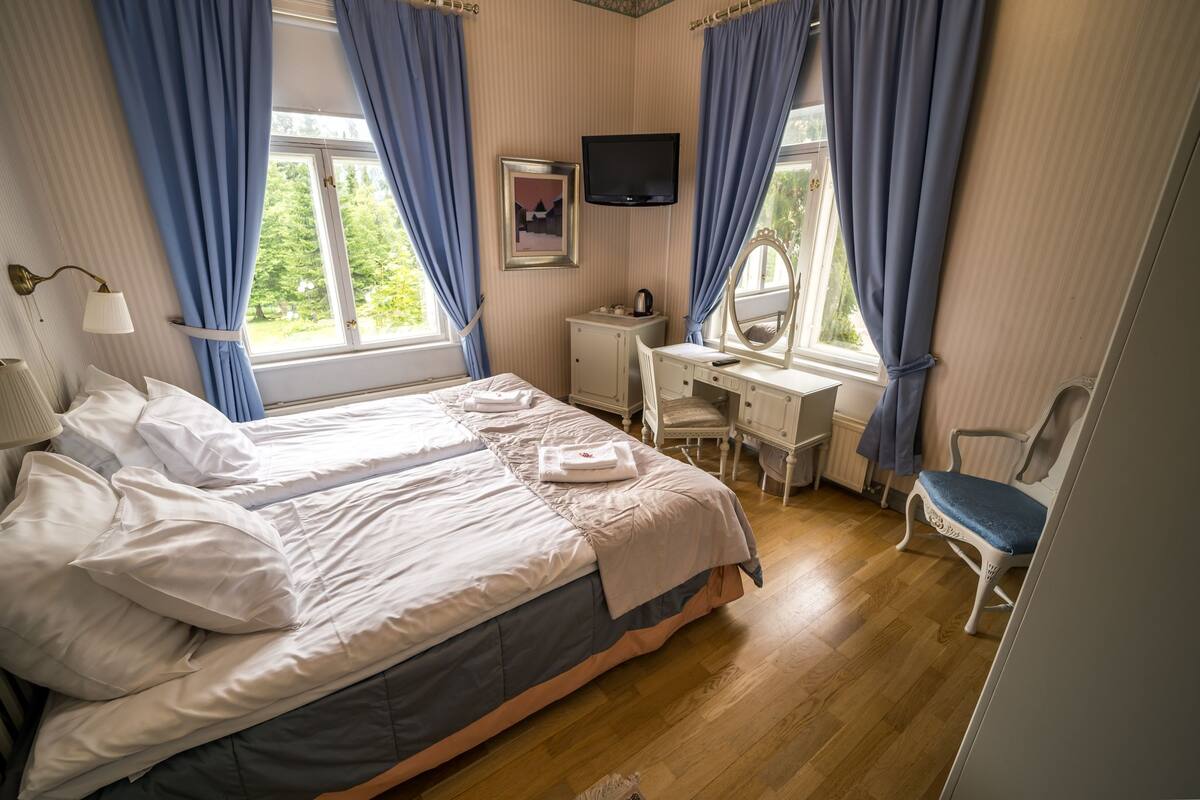 Een net opgemaakt bed met witte lakens, een bureau met computer, een stoel en een televisie aan de muur.