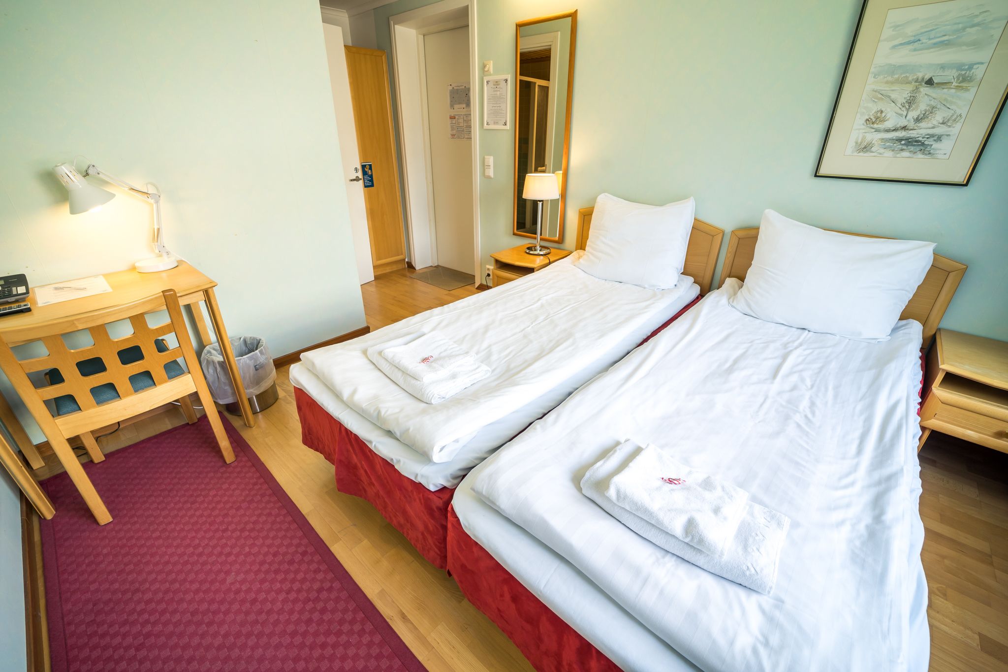 A hotel room with two beds, a desk with a lamp, a chair, and a red carpet.