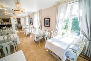 Un comedor con mesas y sillas blancas, candelabros y ventanas amplias con cortinas.