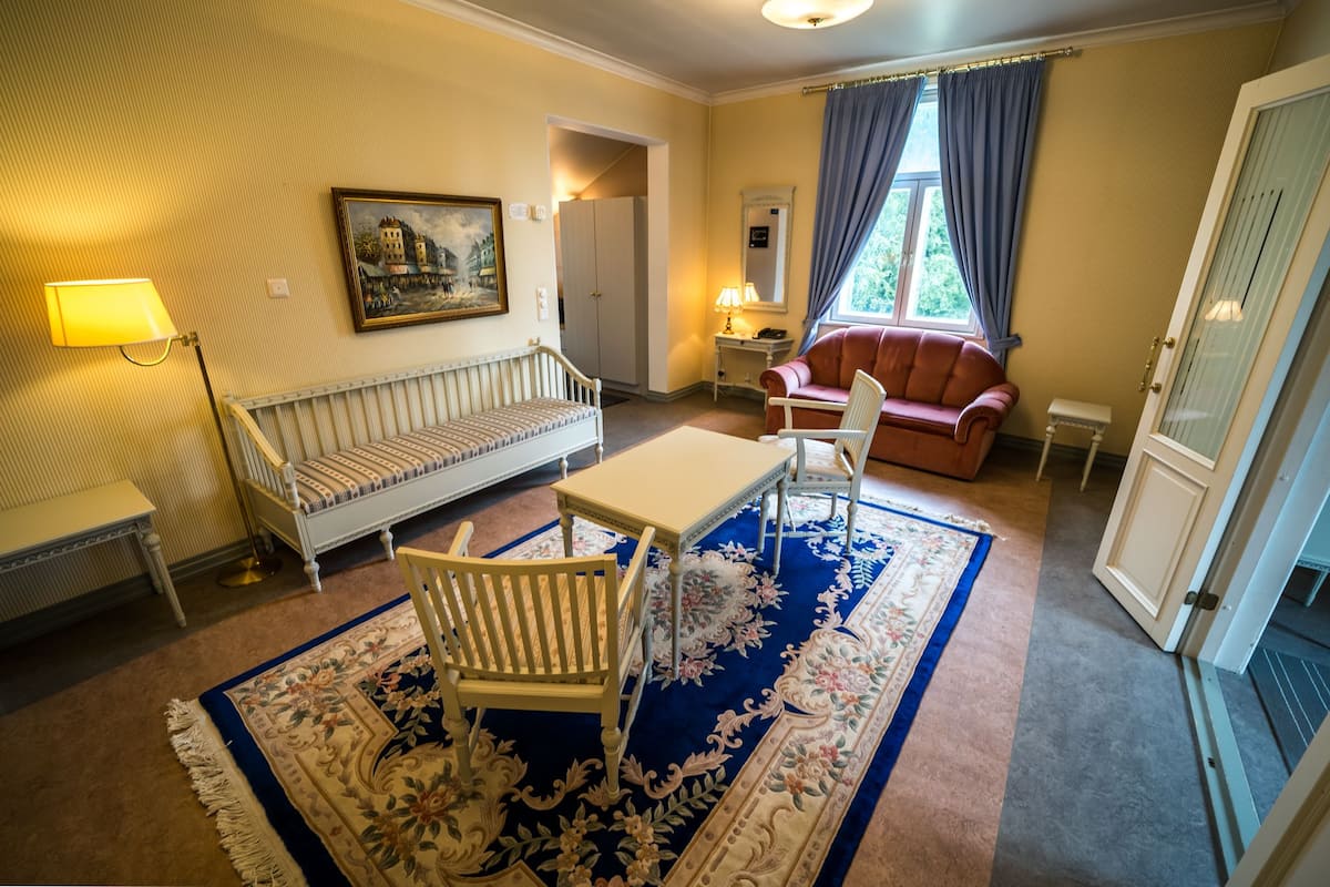 Een kamer met een bank, een tafel, een stoel, een lamp, een schilderij, een raam met gordijnen en een deur.