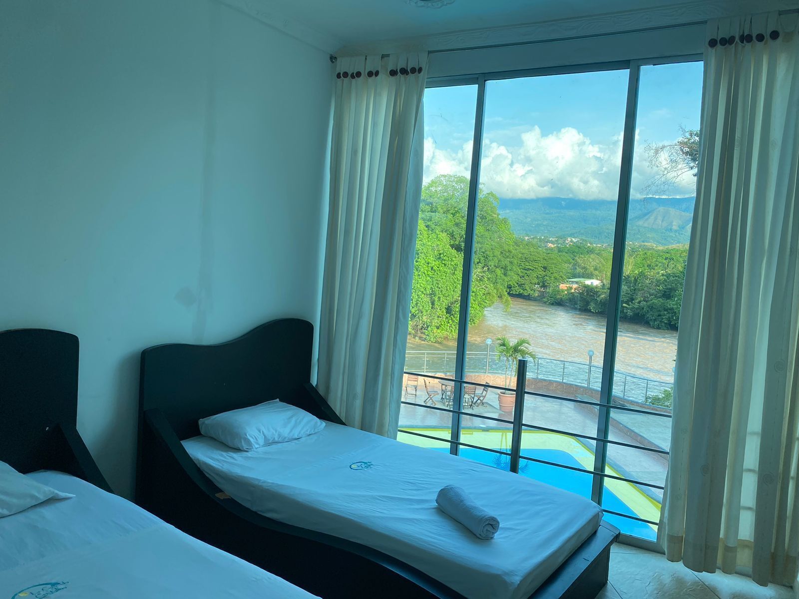 Habitación de hotel con dos camas, un ventanal con vistas al río y vegetación, y un balcón con piscina.