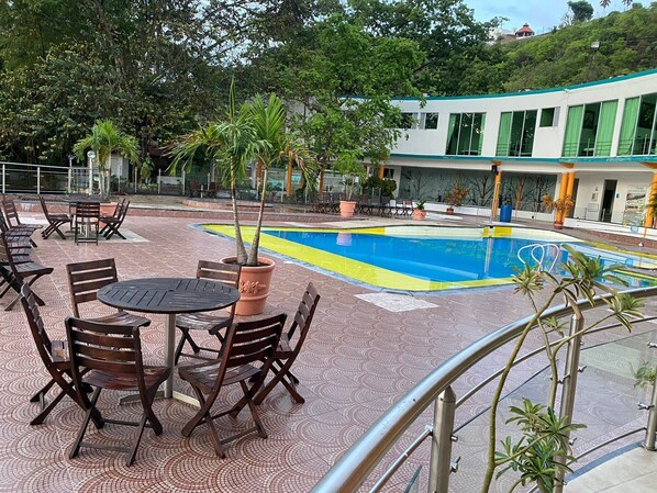 A pool area with a tiled deck, wooden chairs, and a building in the background. - Hotel La Cascada Cristalina (Melgar)