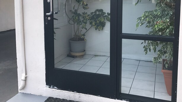 Una puerta de vidrio con marco negro, un letrero que dice "PULL" (TIRAR), una planta en maceta y un letrero en la pared que indica "OFFICE".