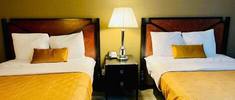 Habitación de hotel con dos camas, cada una con cabecero de madera, ropa de cama blanca y una manta amarilla. Entre las camas, hay una mesita de noche con una lámpara.