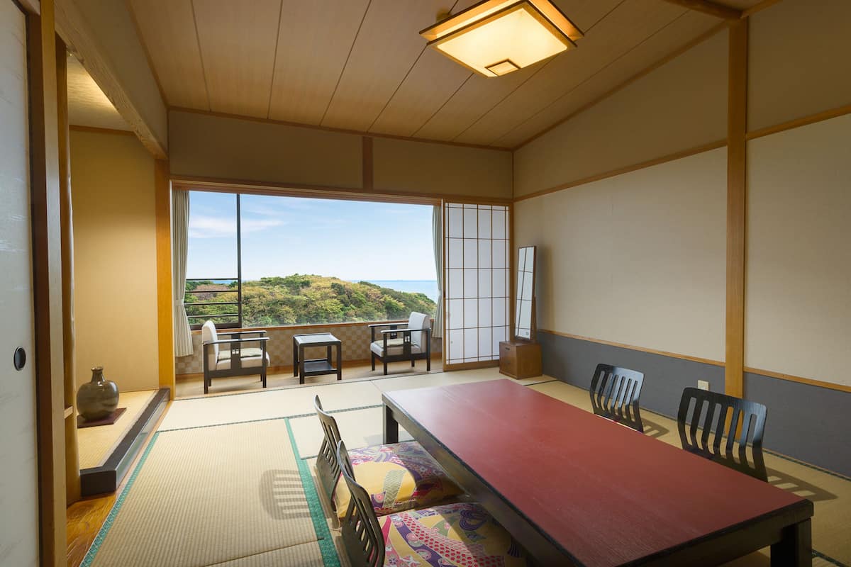 A traditional Japanese room with tatami flooring, a low table, and chairs. A large window offers a view of a hillside.