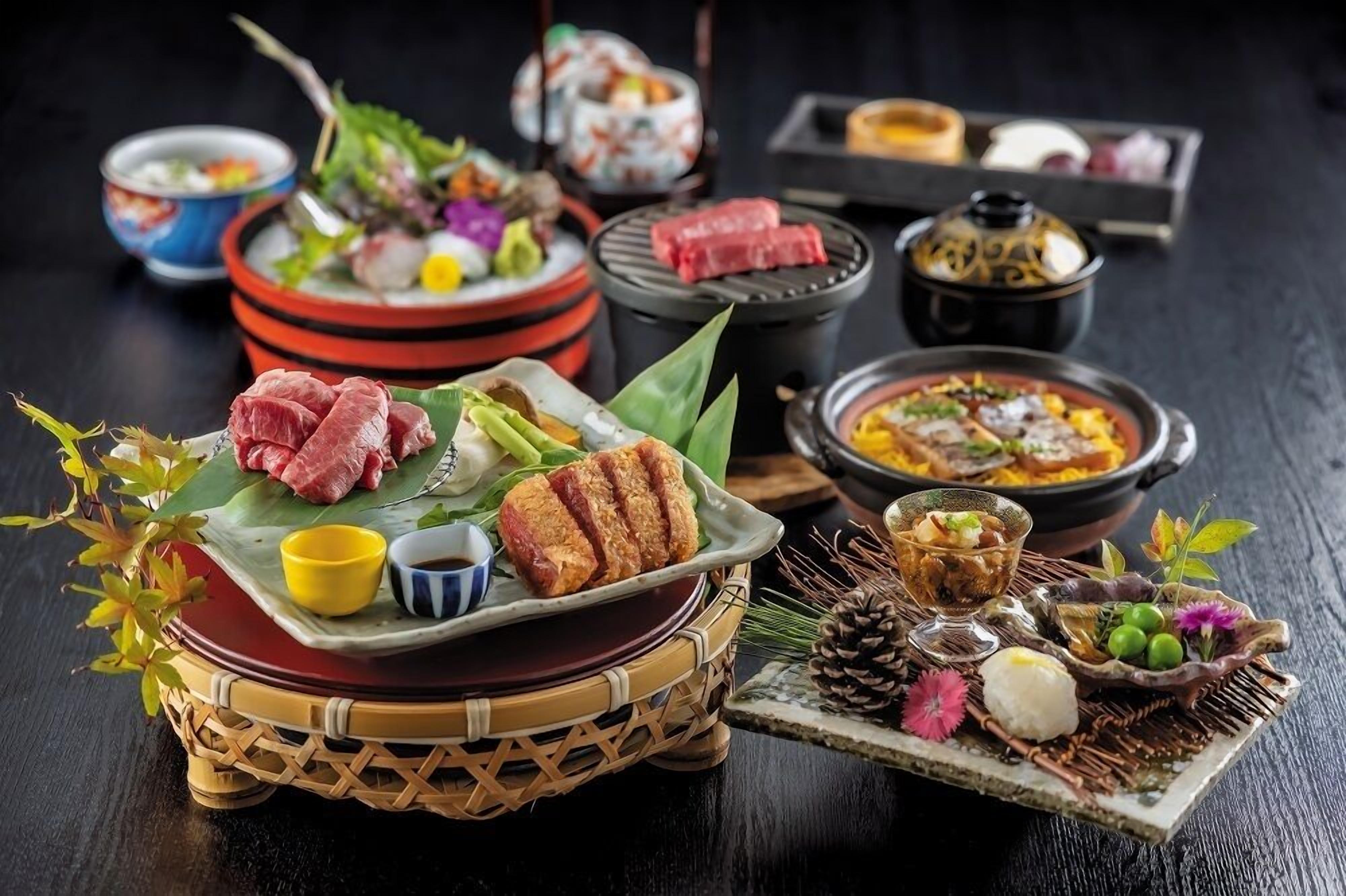 A variety of dishes including grilled meat, sushi, and a bowl of soup, arranged on a table with a black background.